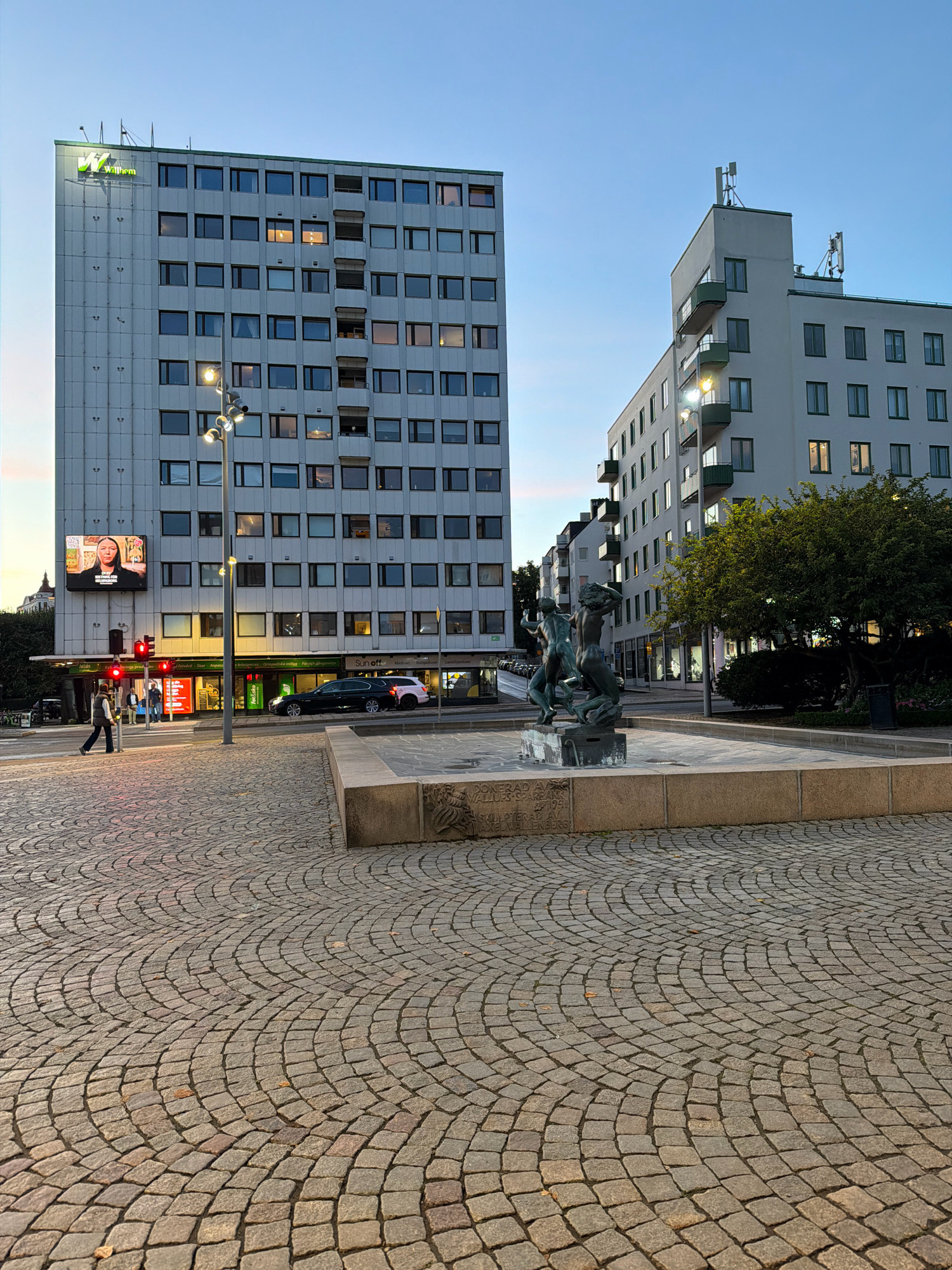 The image captures a street scene in Helsingborg, Sweden, with two modern apartment or office buildings flanking a public square. The building on the left features a prominent green logo at the top, and a large digital billboard displaying a woman's face and text. The building on the right has balconies and visible antennas on its roof. In the foreground, a cobblestone plaza surrounds a low-rise concrete structure, likely a fountain base, with a bronze sculpture of figures on top. The sculpture depicts three human-like forms intertwined. A street lamp with multiple bulbs stands in the middle ground. A person is walking across a street intersection marked by traffic lights, with a black car and a white car parked nearby. The sky is a clear blue, suggesting it is either early morning or late afternoon. The overall impression is one of urban development with historical elements like the sculpture and cobblestone paving.