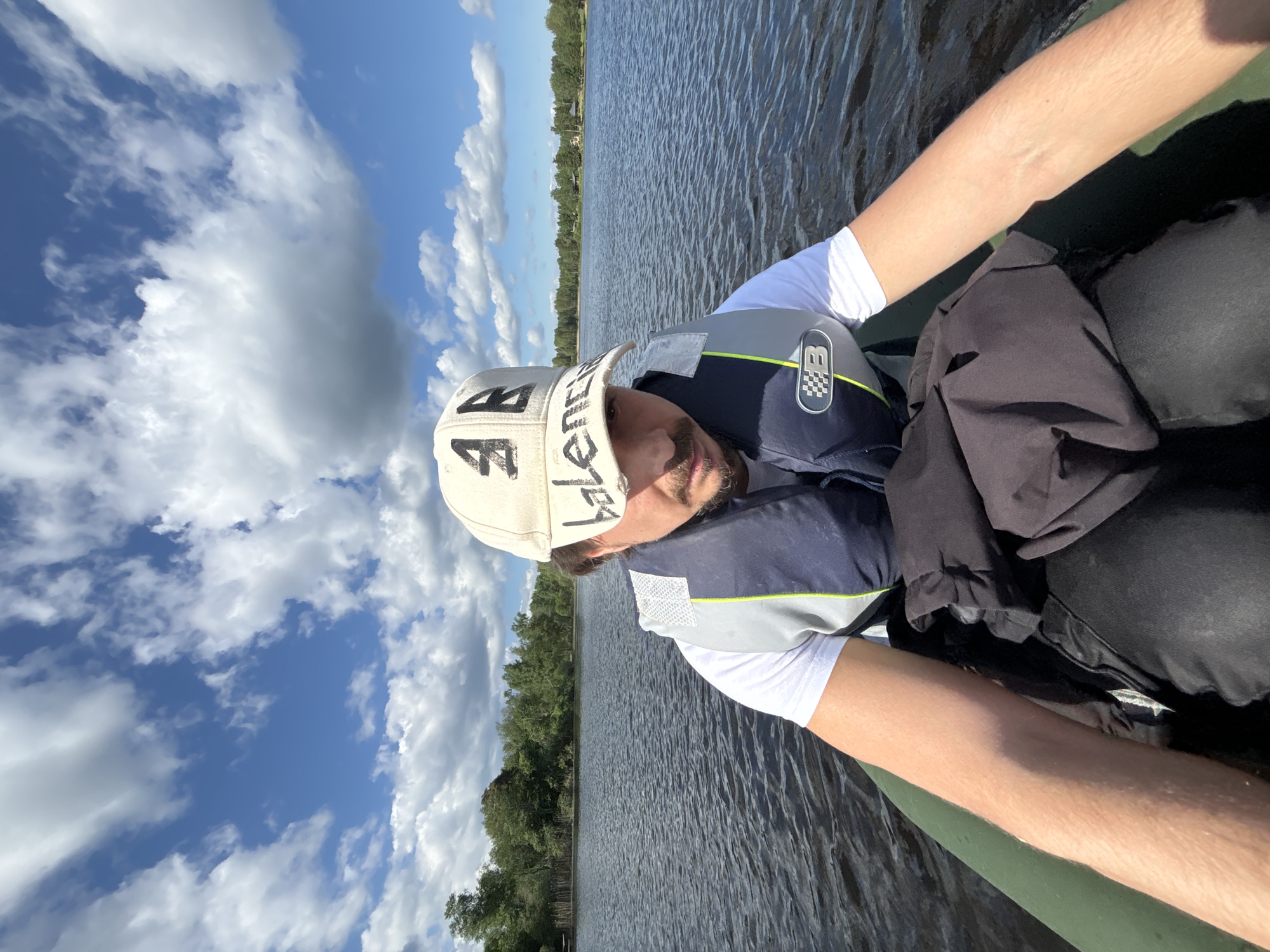 A man in a small boat on a lake. The man is wearing a white t-shirt, a life jacket, and a beige baseball cap with black lettering. He has light skin and a beard. The boat appears to be a kayak or canoe. The lake is calm with trees visible along the shoreline. The sky is blue with scattered clouds. The location is Unknown, Unknown.