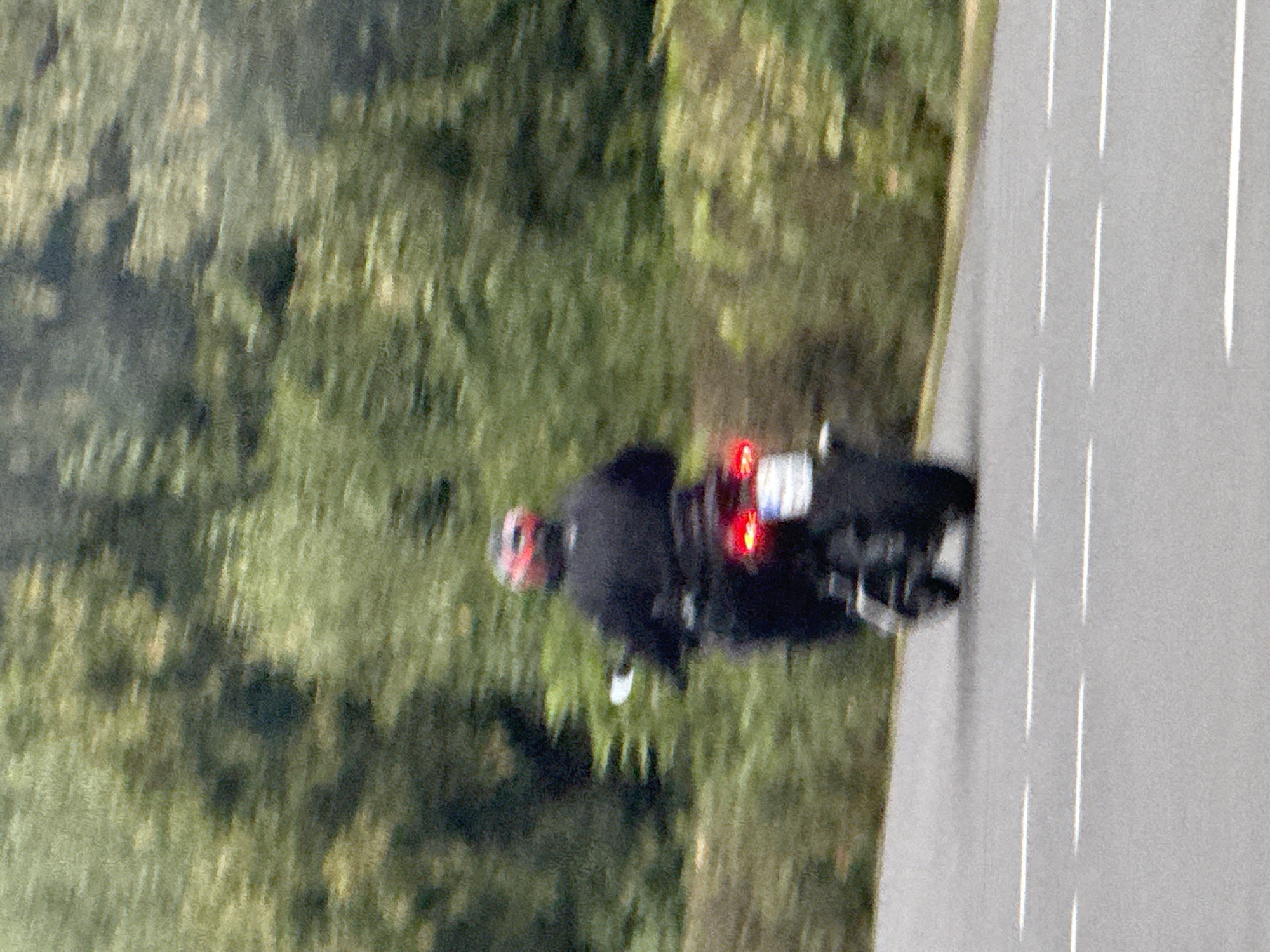 The image shows a motorcycle with a rider from behind, moving away from the viewer on a paved road. The rider is wearing a black jacket and a red helmet. The motorcycle has a lit red taillight and a license plate that is mostly obscured by motion blur. The road has white dashed lines marking the lanes. In the background, there are blurred green trees and bushes. The scene is set in Bad Sachsa, Germany, indicated by the license plate format, which is characteristic of German plates. The image appears to be taken with a long exposure or in low light conditions, resulting in significant motion blur throughout the scene.