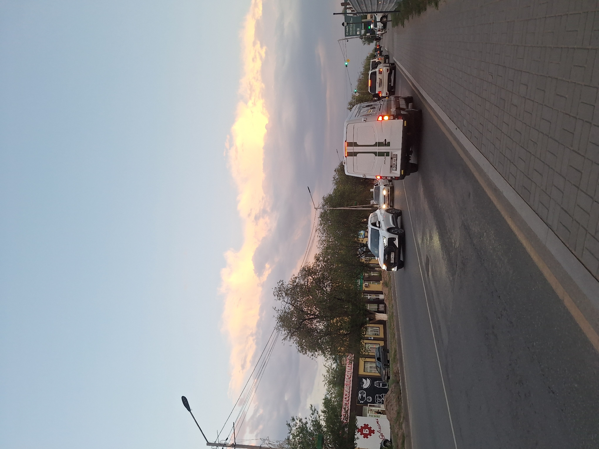 The image shows a street scene in Qonaev, Kazakhstan, with traffic and buildings. The sky is partly cloudy with the sun illuminating the clouds. On the left side of the road, there are low-rise buildings with storefronts, one of which has signage that appears to be in Cyrillic. Several cars and a white van are on the road, moving in the same direction. A pedestrian sidewalk made of paving stones runs along the right side of the road. A street light is visible on the left side of the image. The overall impression is of a street on the outskirts of a city during the late afternoon or early evening.