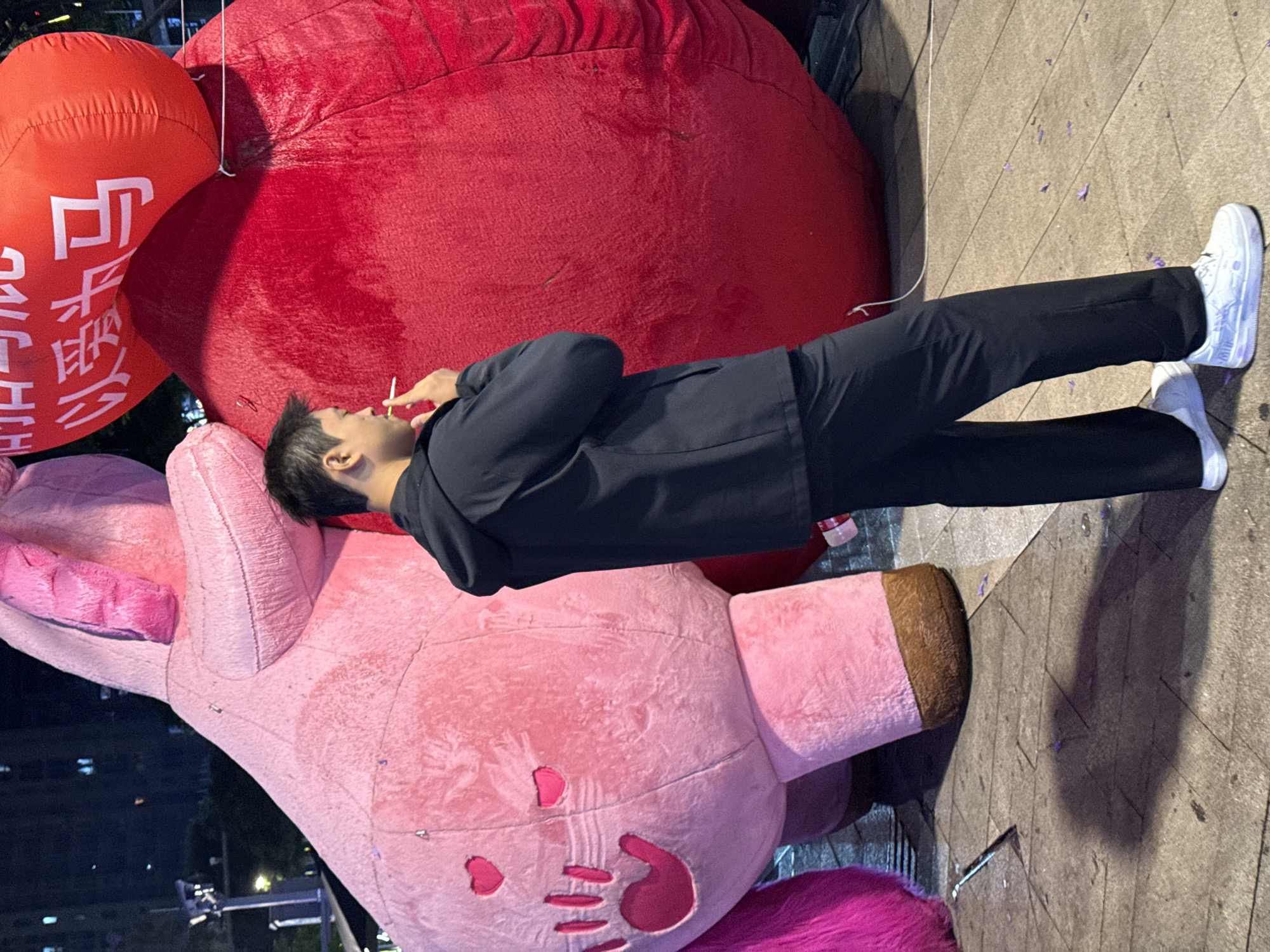 A man is standing near an inflated pink bear and two large, red balloons, one of which features white Chinese characters. He is wearing black pants, a black jacket, and white sneakers. He appears to be smoking. The setting is urban, likely Liangjiang, China, although specific landmarks are not visible in the frame. The scene is situated on a paved surface. Small, violet flower petals litter the ground. Streetlights are visible in the background.