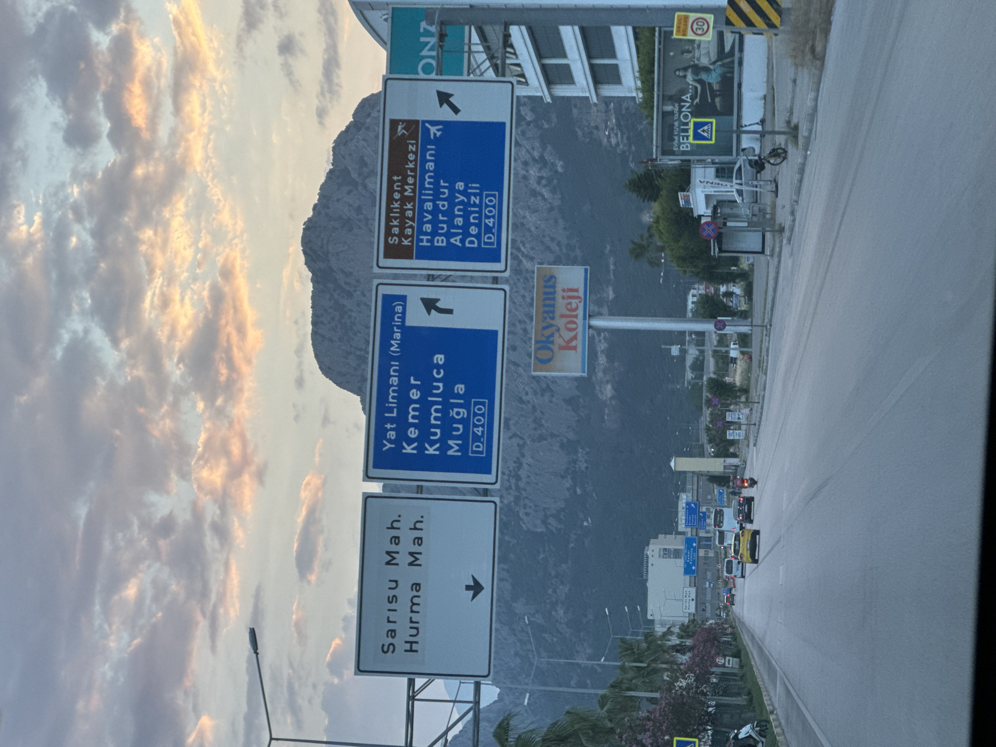 The image presents a roadside view in Konyaaltı, Turkey, likely captured from a moving vehicle, featuring prominent road signs against a backdrop of a majestic mountain and a cloudy sky. The road appears to be a multi-lane highway or main thoroughfare, with several cars, including a yellow taxi, visible further down the asphalt.
A series of large road signs provides directional information. The uppermost blue sign, indicating a right turn, points to "Saklıkent Kayak Merkezi" (Ski Center), "Havalimanı" (Airport), "Burdur," "Alanya," and "Denizli," identifying the route as "D-400." Below it, another blue sign, indicating a left turn, directs to "Yat Limanı (Marina)," "Kemer," "Kumluca," and "Muğla," also on the "D-400" highway. A white sign below these points right to local neighborhoods: "Sarısu Mah." and "Hurma Mah." Further down the road, a blue sign for "Okyanus Koleji" (Okyanus College) is visible.
The background is dominated by a rugged, rocky mountain, its upper slopes partially obscured by the sky. The sky is filled with clouds, some of which are illuminated with warm, soft light, suggesting either sunrise or sunset. Urban elements line the right side of the road, including buildings, streetlights, and a bus stop. An advertisement for "Bellona" furniture, displaying "EVİMİ YUVA YAPAN BELLONA," is seen, along with a "30" km/h speed limit sign and a pedestrian crossing sign. The scene captures a busy transportation artery set against a dramatic natural landscape.
