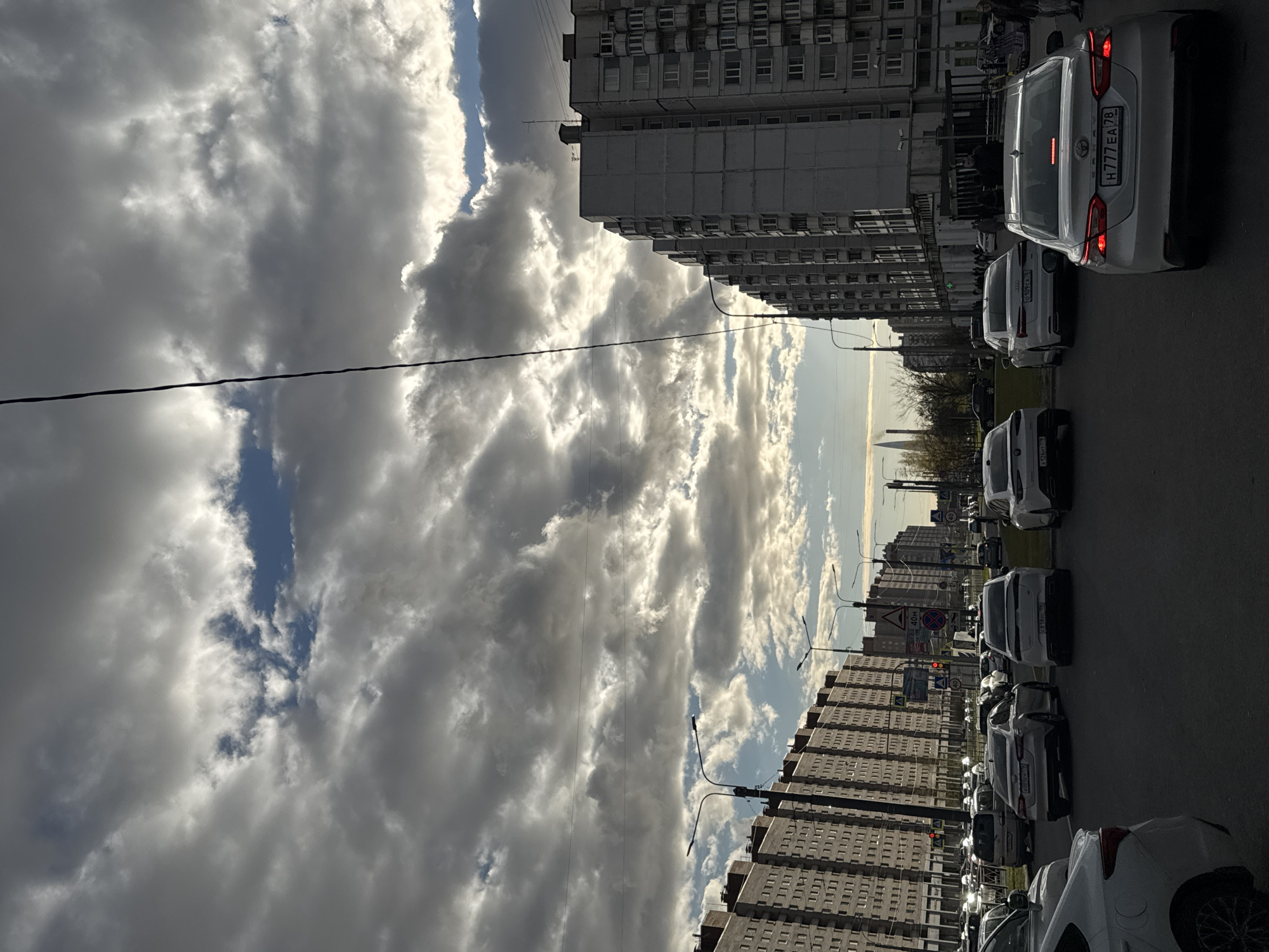 The image captures an urban street scene in Saint Petersburg, Russia, under a dramatic, partly cloudy sky. Large, textured cumulus clouds, varying from bright white to dark grey, dominate the upper and middle portions of the frame, with visible patches of blue sky and bright sunlight breaking through. A black wire or power line stretches horizontally across the sky.
On the right, a long line of multi-story apartment buildings, indicative of Soviet-era architecture, borders the street. These grey structures feature numerous windows and balconies or air conditioning units, creating a uniform, repetitive pattern. Along the street parallel to these buildings, several cars, mostly white or light-colored sedans, are either parked or moving slowly in a line. Street lights with distinctive, curved arms extend upwards from the side of the road.
Visible text on the cars' license plates confirms the location. A white Toyota sedan in the foreground clearly displays the Russian plate "H777 EA 78". Another white Audi behind it shows "009 EA 178". The "78" and "178" are regional codes for Saint Petersburg. Other visible details include a road sign with "40m" (likely indicating distance) and a red circular road sign further down the street.
The time of day appears to be daytime, possibly late afternoon or early evening, characterized by the bright, angled sunlight creating strong contrasts and shadows. The weather is dynamic, with significant cloud cover but also clear sky, suggesting a changing atmospheric condition. The scene portrays a typical bustling urban environment with characteristic Russian architecture and street activity.