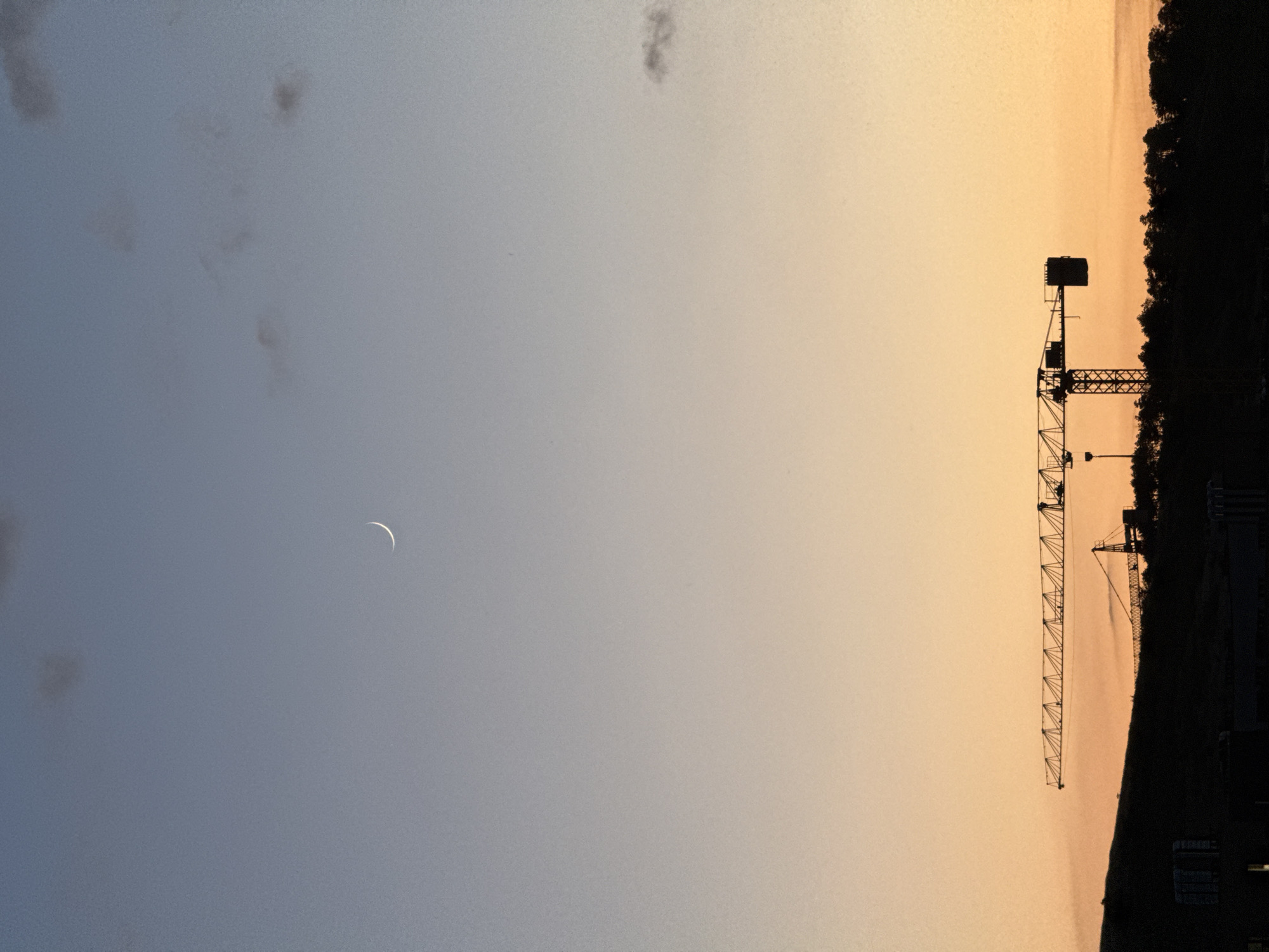 The image captures a twilight scene in Burgas, Bulgaria, featuring a silhouetted construction crane against a gradient sky of blue to orange. A thin crescent moon is visible in the upper portion of the sky. The lower half of the image is dominated by the dark silhouette of a landscape, which includes the distinct structure of a large construction crane stretching horizontally across the frame. A second, smaller crane is also visible behind a tree line. The overall impression is one of urban development against a natural backdrop.