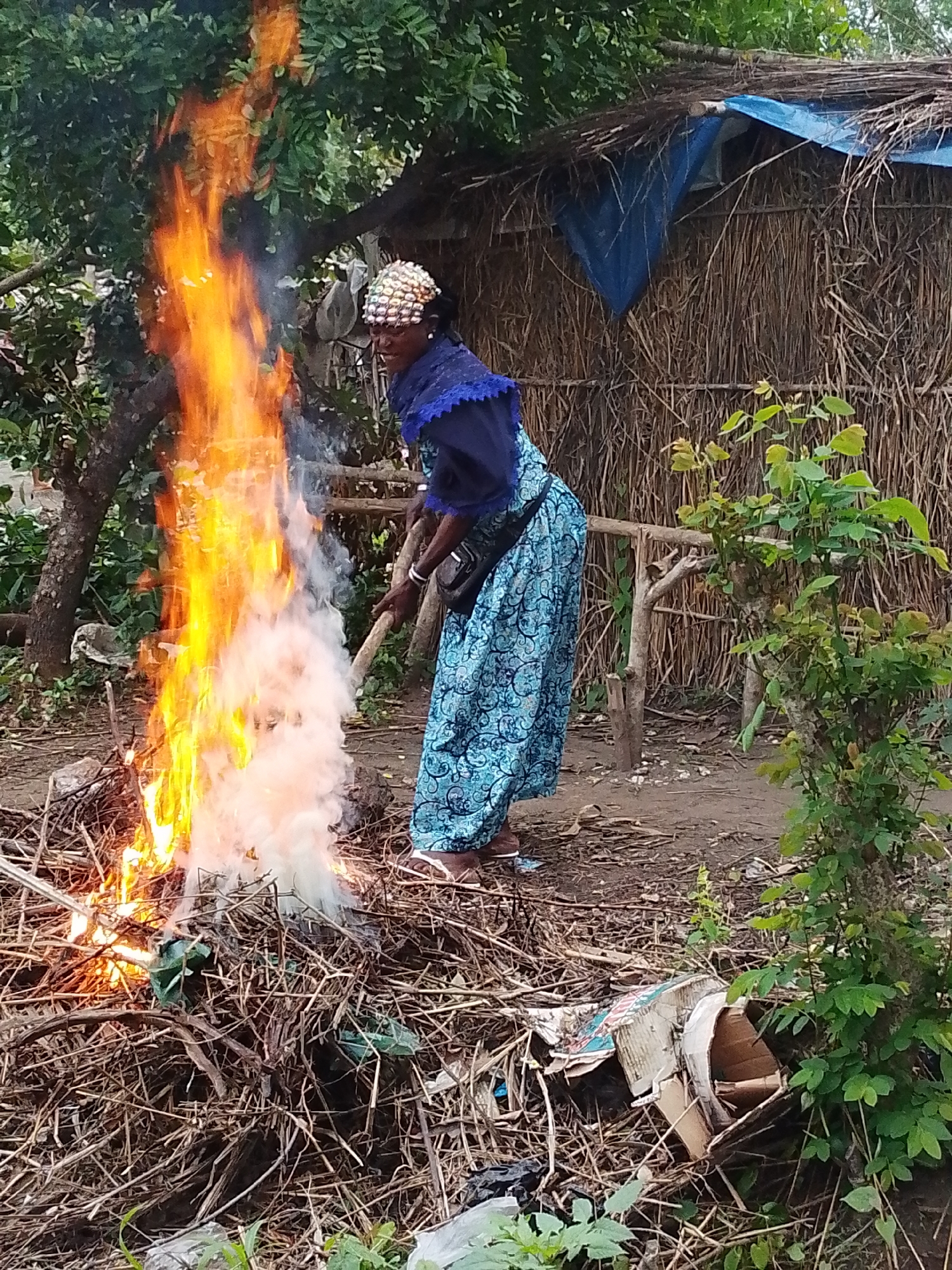 An individual, appearing to be a woman wearing a blue patterned dress and a beaded head covering, is standing by a large, roaring bonfire of dried plant material and debris. She is holding a long stick, potentially to tend the fire. Behind her, a rustic structure, possibly a dwelling, is visible, constructed with thatch and adorned with a blue tarp. The environment appears to be rural, with trees and vegetation surrounding the scene. The ground is dirt and littered with more dried plant matter and some discarded cardboard boxes. The scene is set in Otulu, Nigeria.