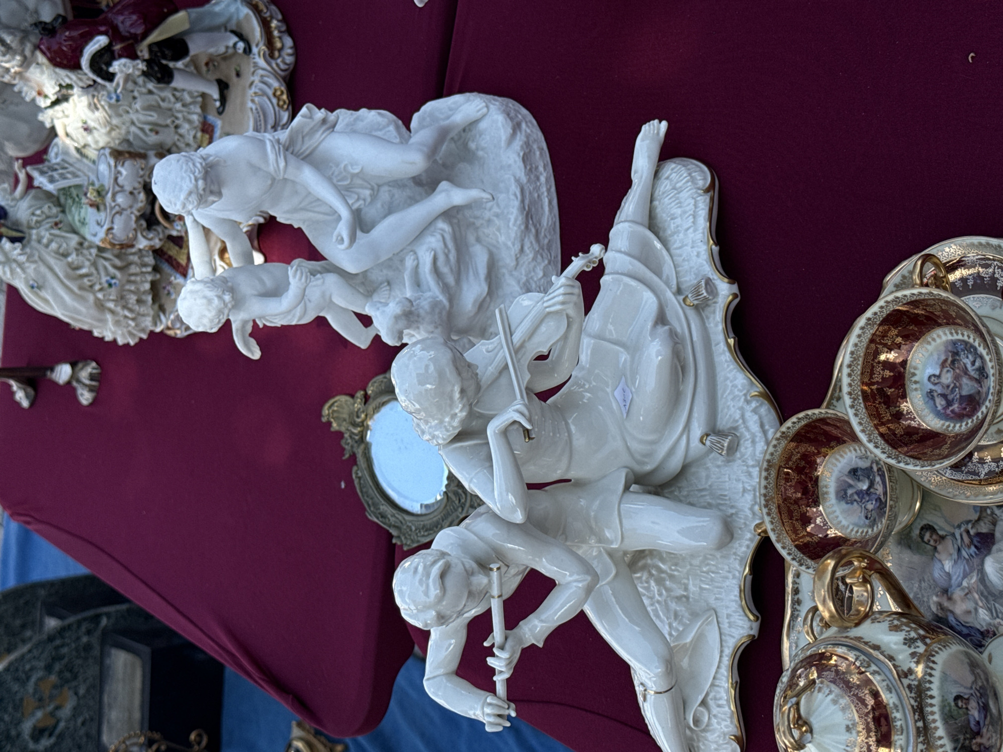 The image displays a collection of porcelain figurines and a tea set arranged on a deep red tablecloth. In the foreground, a white porcelain figurine depicts a male figure playing a flute and a female figure playing a violin, positioned on a decorative base. Behind them, another white porcelain figurine shows a woman with a child. Further in the background, a more elaborate porcelain scene with multiple figures and ornate details is visible. A vintage mirror with an ornate frame sits behind the musical figurines. In the bottom portion of the image, a porcelain tea set with gold trim and intricate painted scenes is displayed. The scene appears to be set up at an outdoor market or antique fair. The setting of Bruges, Belgium is not evident from the image itself.