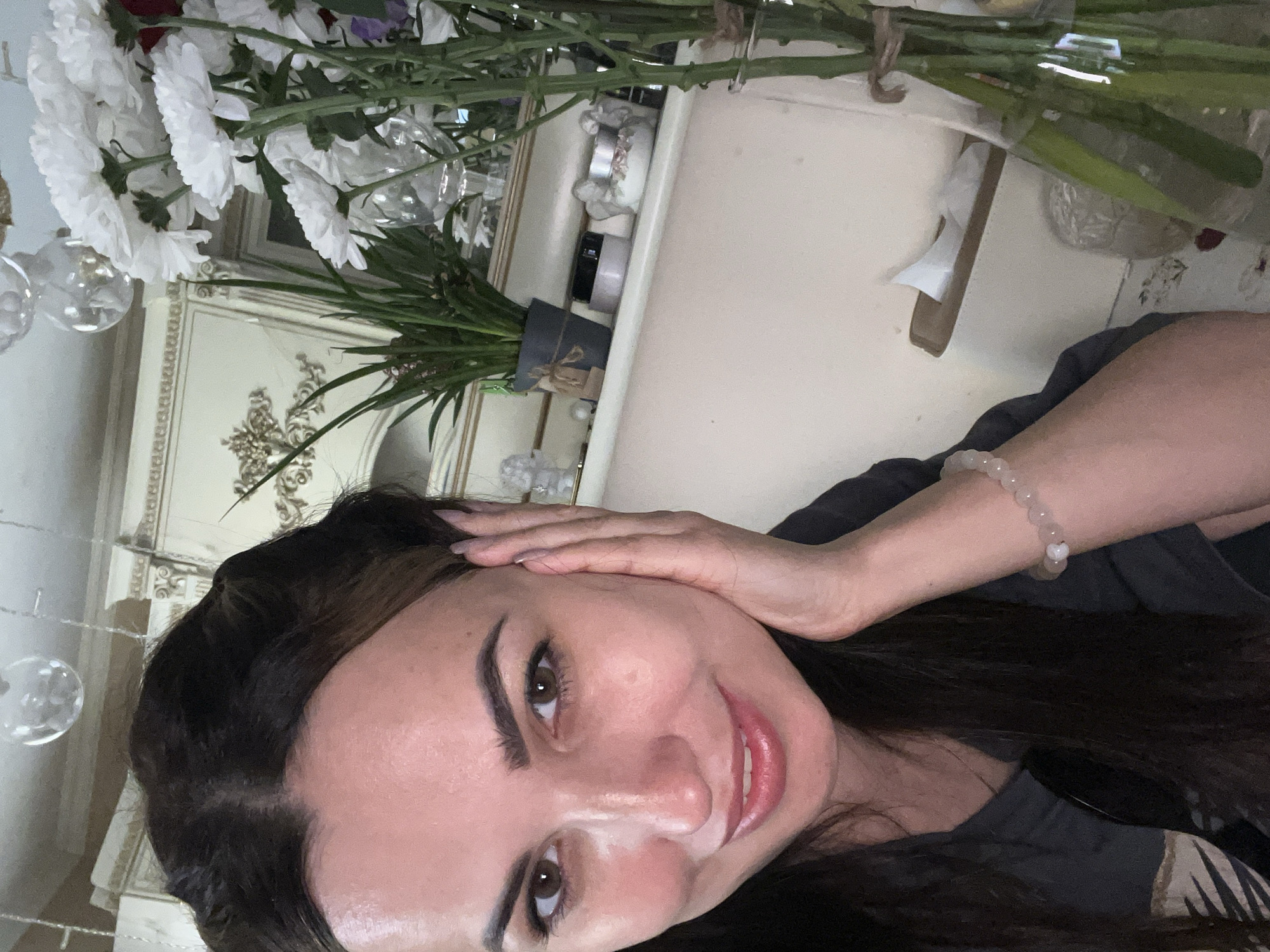 A medium shot captures a woman posing for a selfie in what appears to be an indoor setting, possibly a home or studio. She has long, dark hair styled with some volume around her temples and is resting her right hand on the left side of her face. Her eyes are open, and she is smiling gently with her lips slightly parted. She is wearing a dark-colored t-shirt and a beaded bracelet on her right wrist. In the background, to the right of the woman, a bouquet of white chrysanthemums in a clear glass vase is visible. The vase is tied with twine. Further in the background, a white shelf holds a potted plant, a jar, and decorative figurines, with elaborate cabinetry and decorative molding on the wall behind it. The lighting suggests an indoor environment. There is no information available to determine the location of the city or country.