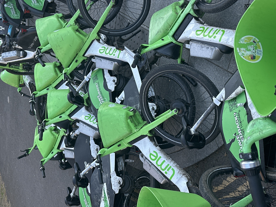The image displays a significant, disorganised accumulation of approximately two dozen electric bicycles, predominantly colored neon green and white. Each bicycle is prominently branded with the "Lime" logo, and many units also feature "Lime + Uber" branding along with QR codes labeled "HAVE FEEDBACK? SCAN HERE TO SHARE." The e-bikes are haphazardly stacked and interlocked, with numerous units overturned, resting on their sides, or tangled within the pile. Visible wear includes dirt, scuff marks, and peeling decals on the frames, handlebars, and front baskets. The scene is set on a grey, paved surface, indicative of an urban public space such as a sidewalk or plaza, located within the city of London, United Kingdom, where Lime e-bike services operate. No individuals or other discernible objects are present in the immediate vicinity.