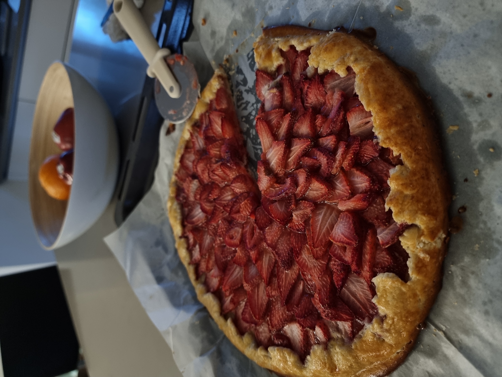 The image displays a close-up, slightly tilted view of a freshly baked strawberry galette or free-form tart, positioned on a kitchen counter.
The central subject is a roundish galette with a golden-brown, rustic crust that is visibly folded over the edges of the fruit filling. A large, wedge-shaped slice has been removed from the galette, revealing the densely packed filling of vibrant red, thinly sliced strawberries. The strawberries appear juicy and have a slight sheen, possibly from their natural juices or a glaze. The remaining crust is flaky and irregular, characteristic of a homemade rustic tart. The galette rests on a sheet of crumpled white or light grey parchment paper, which is spread over a light-colored surface, likely a kitchen countertop.
In the background to the left, a light-colored, possibly wooden or ceramic, bowl is visible. Inside the bowl, there are several pieces of fruit; at least one is a bright orange citrus fruit, resembling an orange or tangerine, and another is a darker, reddish-orange fruit. Above and slightly behind the bowl, a pizza cutter is hanging or resting. It features a light cream or beige plastic handle and a grey circular blade with some reddish residue on it. Parts of a dark object, possibly a cutting board or another utensil, are also faintly visible behind the bowl and pizza cutter. Further back on the left, a reflective, metallic surface, likely stainless steel (perhaps an appliance or backsplash), catches some light. The overall setting suggests a domestic kitchen environment.
The image captures a moment related to baking and enjoyment, with the galette having just been prepared or served, as indicated by the missing slice. The lighting appears to be indoor, perhaps natural light or soft artificial light, illuminating the food and creating a warm ambiance. No text is visible in the image.
