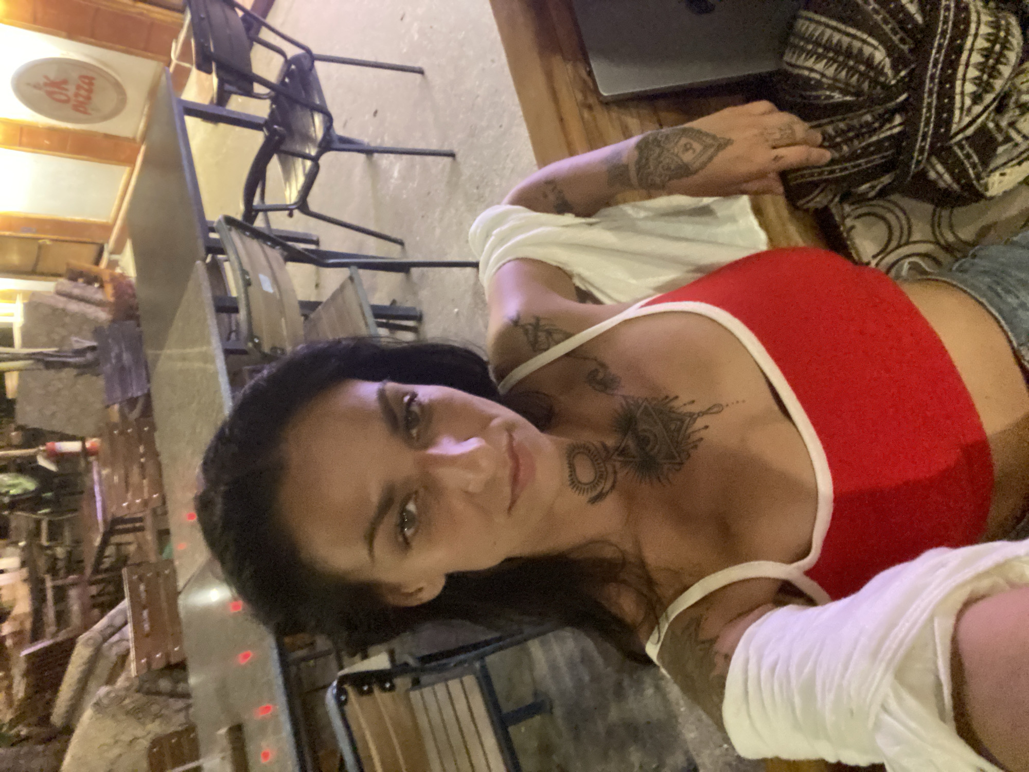 A woman with dark hair and tattoos on her arms and chest is taking a selfie in what appears to be an outdoor seating area of a restaurant. She is wearing a red tank top with white trim and has a white shirt draped over her shoulders. In the background, there are tables and chairs, some of which appear to be made of wood. A sign with "OK PIZZA" is visible on a wall in the distance. The ground is concrete, and there is a patterned fabric on a seat next to the woman. The lighting suggests it is evening or night. The location is identified as Phu Thuy Ward, Vietnam.