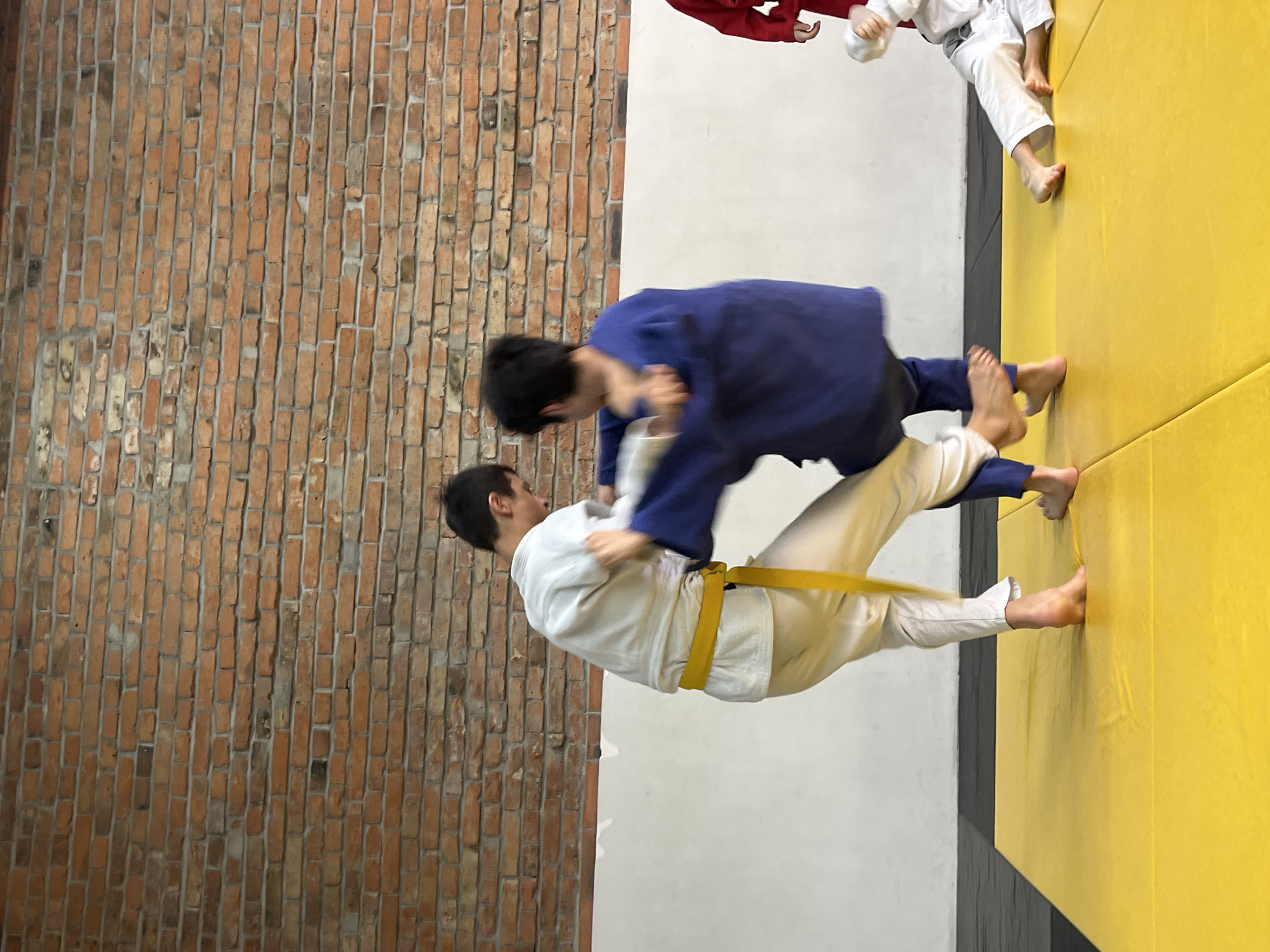 Two people are engaged in a judo-style grappling activity on a yellow mat. One person is wearing a white gi with a yellow belt, while the other wears a blue gi. They are in the midst of a throw or takedown maneuver.
The background consists of a brick wall on one side and a plain wall. Additional people wearing gis are partially visible on the periphery of the mat. Based on reverse image searches, this photo was taken in Krasnoyarsk, Russia.