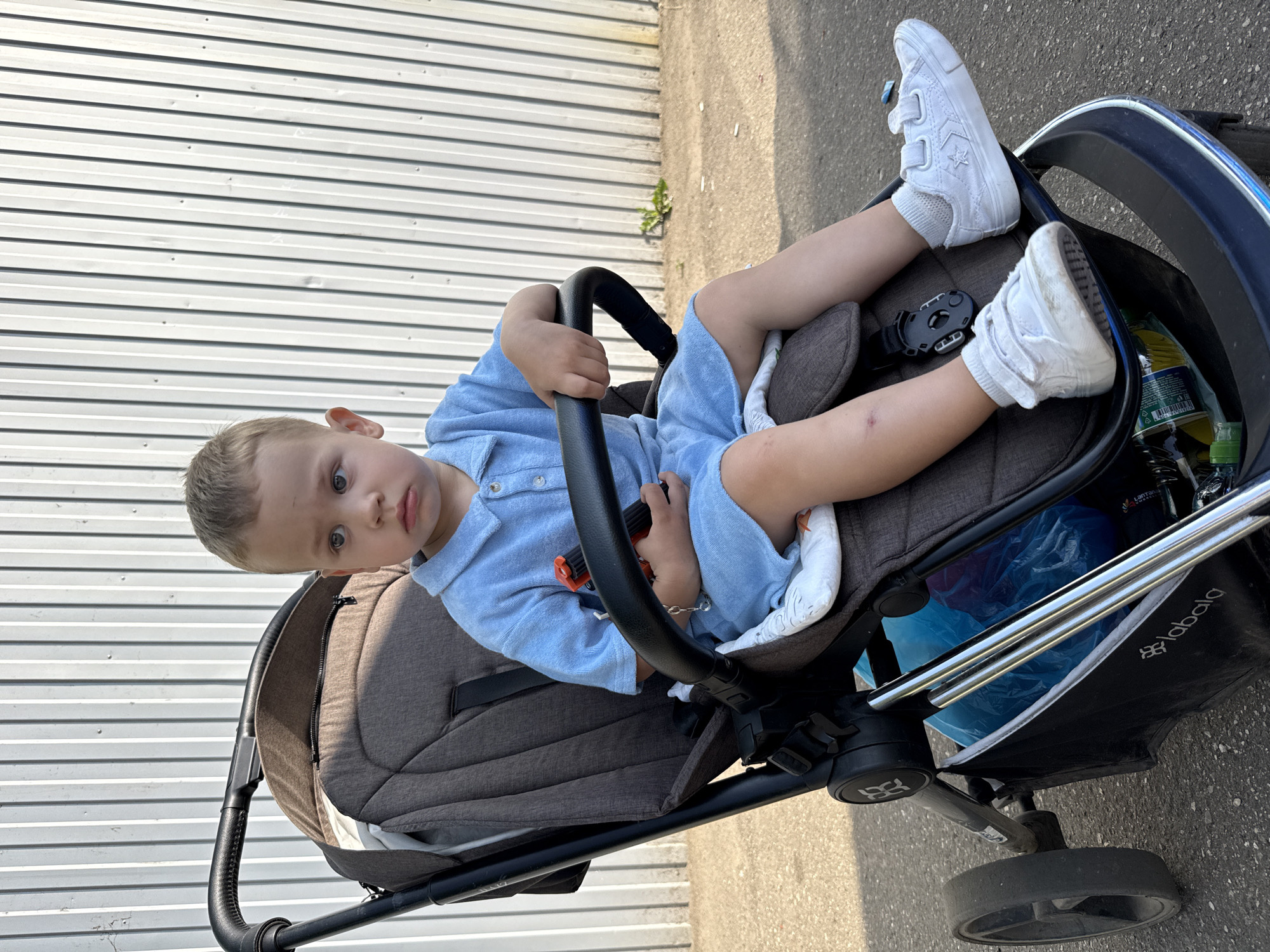 A young boy with light brown hair and blue eyes sits in a stroller, looking to his left with a neutral expression. He is wearing a light blue terrycloth polo shirt and shorts, white socks, and white sneakers. The boy is holding an orange toy in his right hand. The stroller is dark gray and black with chrome accents. A blue plastic bag and a green bottle are visible in the storage compartment beneath the boy. The background is a corrugated metal fence, and the ground is paved asphalt. This image was captured outdoors on a sunny day. The location is identified as Rusanovo, Russia.