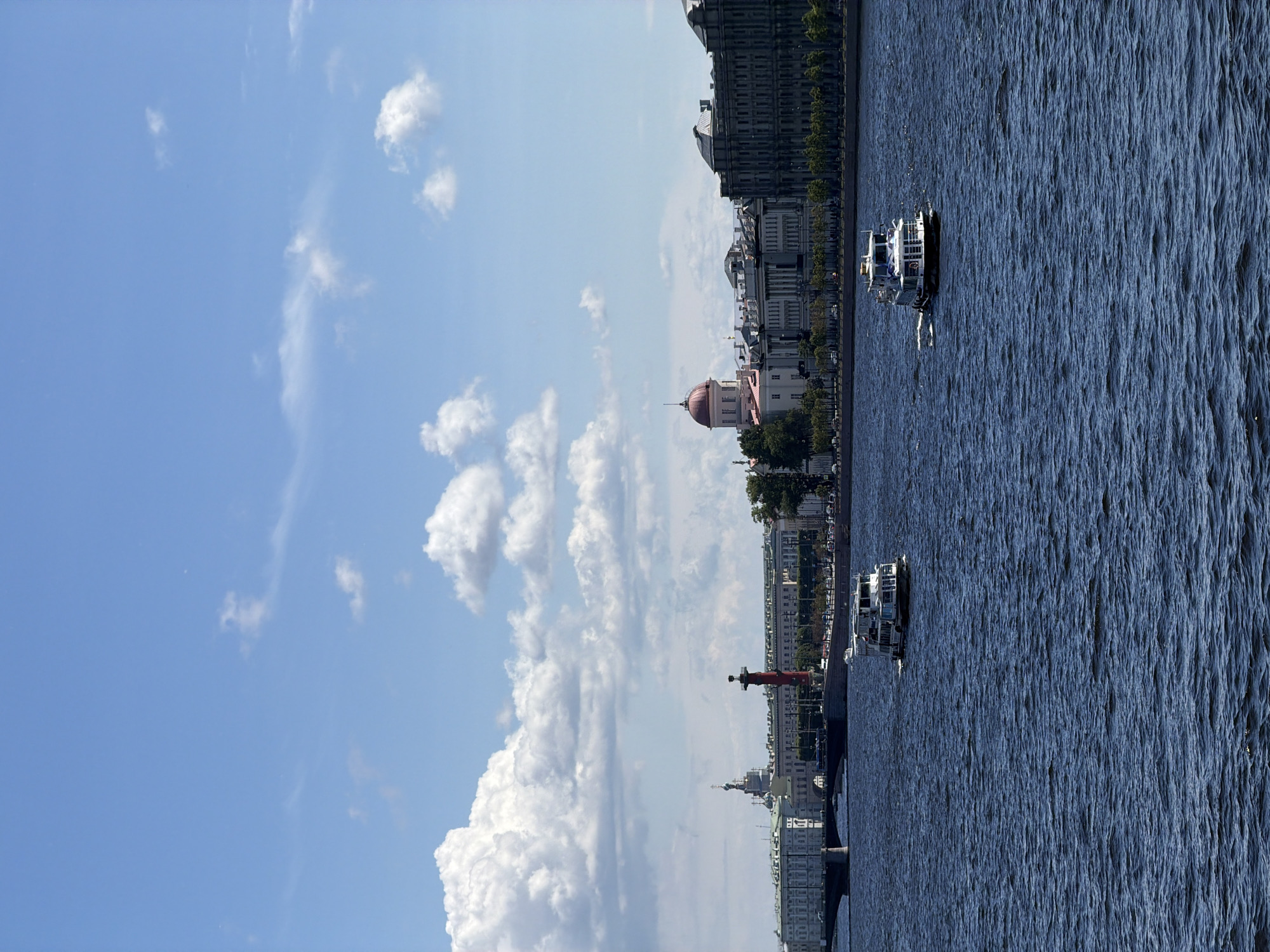 The image captures a wide, blue river in Saint Petersburg, Russia, on a clear day with scattered white clouds. Two white passenger boats are visible on the water. The left boat is farther away, while the right boat is closer to the foreground. Along the riverbank, a long stretch of historical buildings forms the cityscape. Notable structures include a prominent red rostral column with a lantern, a building with a pink dome, and several other classical-style buildings with grand facades. A bridge with arches spans the river in the distance. Trees line the riverbank in front of the buildings, and a few cars are discernible on the road behind them. The overall scene depicts a tranquil, sunny day in a picturesque city.