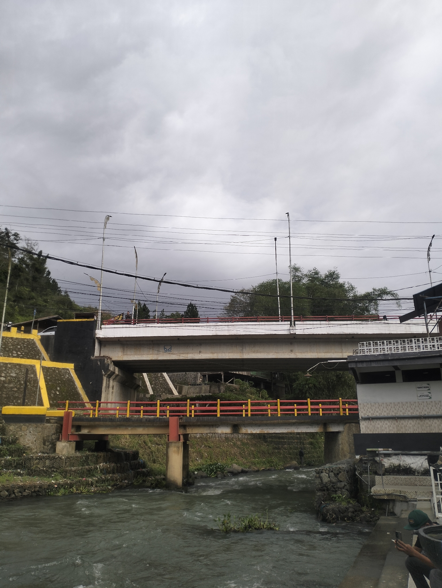 The image depicts a scene in the city of Blang Bebangka, Indonesia, featuring two bridges crossing a river. The foreground shows a murky, fast-flowing river with some aquatic vegetation. To the right of the river, a person wearing a green cap is seated, holding a phone and a cigarette. Adjacent to the riverbank is a structure with a tiled facade and a sign that reads "TEMPAT WUDHU" (ablution area).
A smaller, older bridge with red and yellow railings spans the river closer to the viewer. This bridge is supported by concrete pillars. Behind this bridge, a larger, more modern bridge with a white concrete surface is visible. This bridge also has red railings and is supported by substantial concrete piers. The sides of the embankment leading up to the larger bridge are reinforced with stone and yellow-painted concrete. Power lines crisscross the sky above the bridges. Trees and foliage are present on the hillsides in the background, and the sky is overcast with grey clouds.