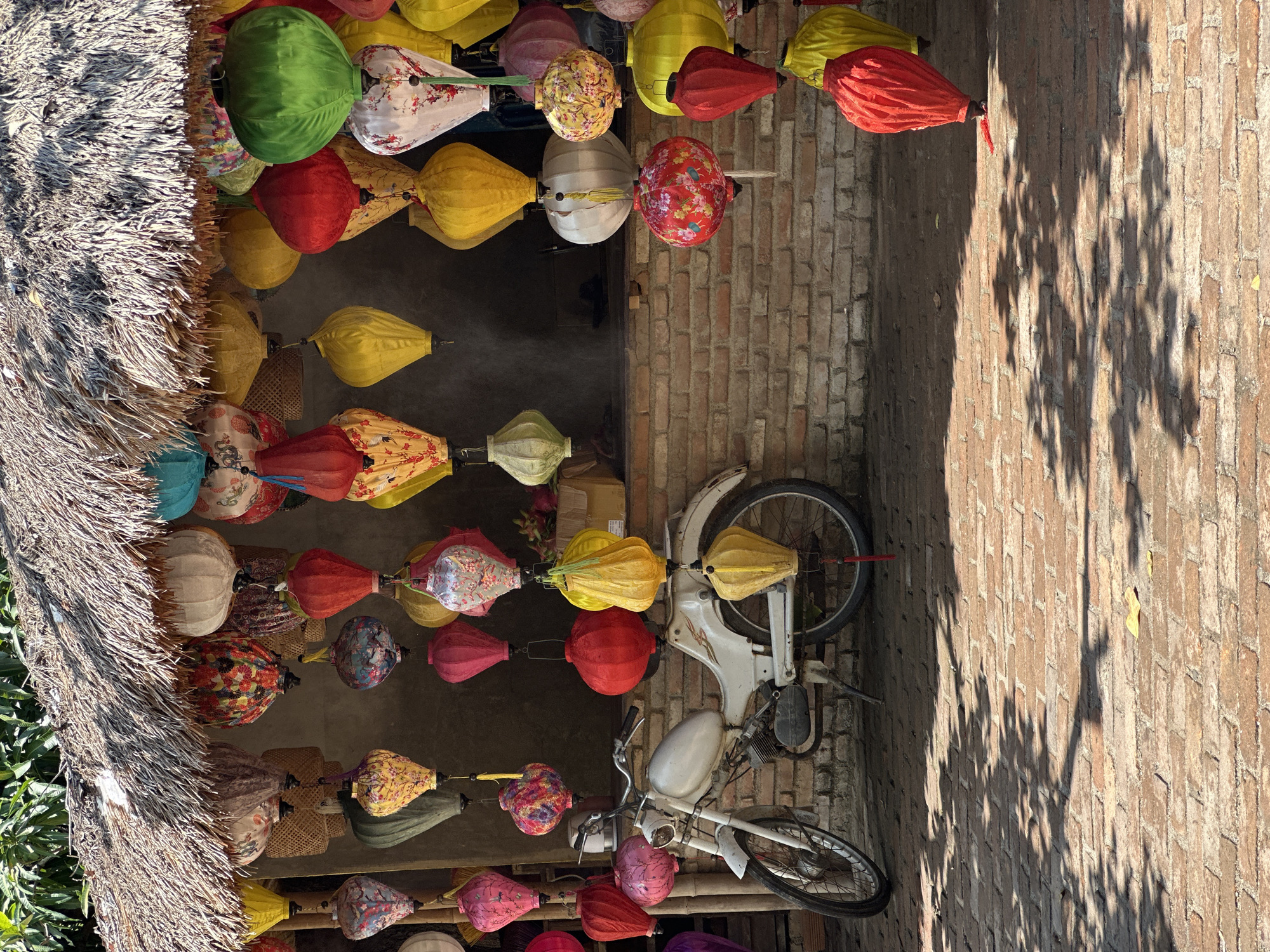 The image depicts a street scene in Nha Trang, Vietnam, featuring a thatched-roof stall overflowing with colorful, traditional lanterns. A white moped is parked in front of the stall, leaning on its kickstand. The lanterns, made of silk and paper, are in various shapes and sizes, with intricate patterns and designs. They hang from the stall's eaves and are arranged in rows, creating a vibrant and festive atmosphere. The stall itself is constructed with rough brick walls and a thatched roof, suggesting a rustic and local establishment. The ground is paved with red bricks, and dappled sunlight casts shadows of leaves from overhead trees onto the pavement. The scene appears to be outdoors, likely in a market or along a street, given the context of a stall selling goods. There are no people visible in the image. The overall impression is one of cultural charm and local commerce.