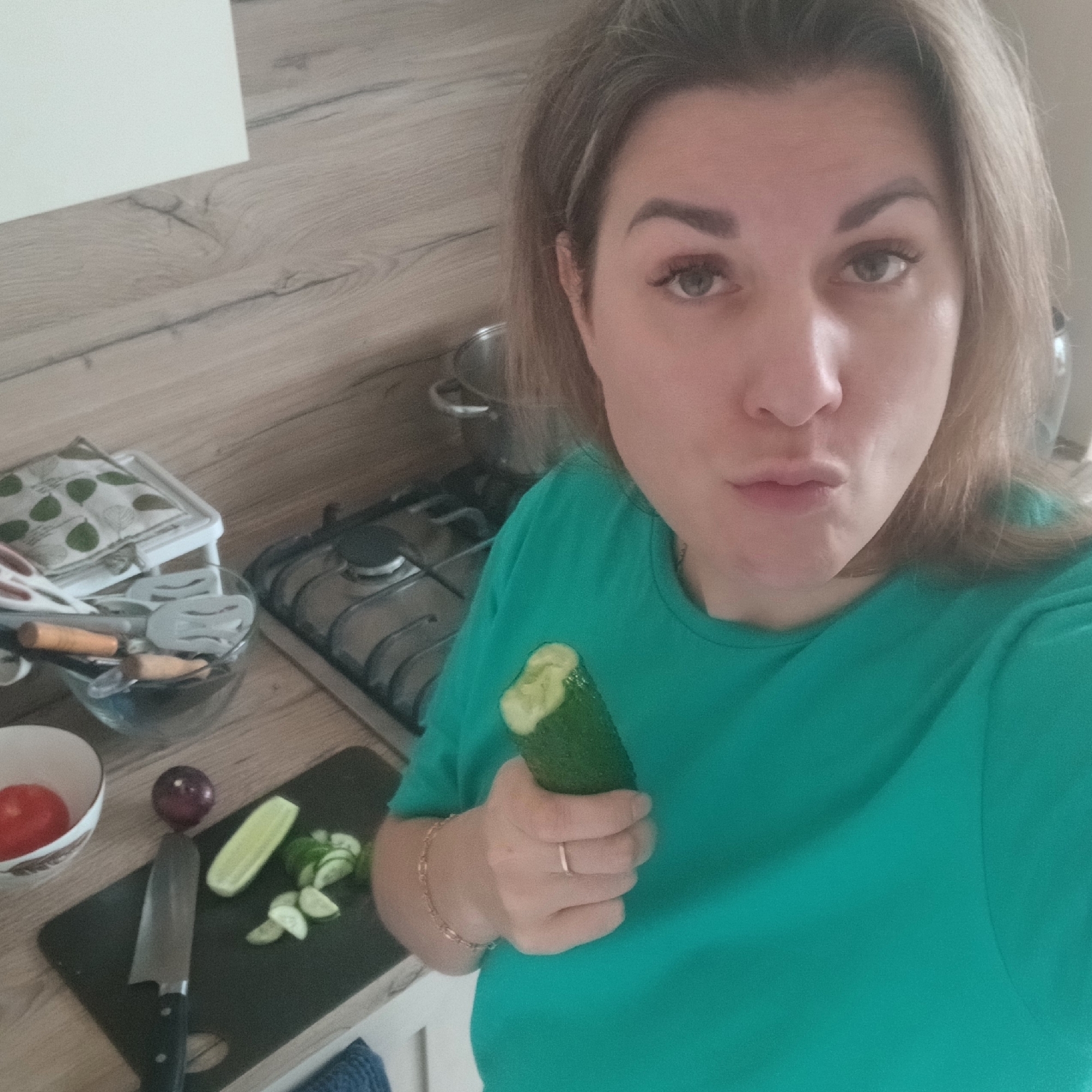A woman with light brown hair is taking a selfie in what appears to be a kitchen. She is wearing a bright green t-shirt. She holds a partially eaten cucumber in her right hand, and her lips are pursed in a kissing expression. On the counter to her left is a black cutting board with sliced cucumber, half a cucumber, an onion, and a large kitchen knife. Behind the cutting board is a white bowl containing a tomato. Further behind, there is a gas stove with pots and pans. There's a glass bowl with kitchen utensils. The countertop has a light brown wood grain pattern. The city of Izhevsk, Russia, is not identifiable in this image.
