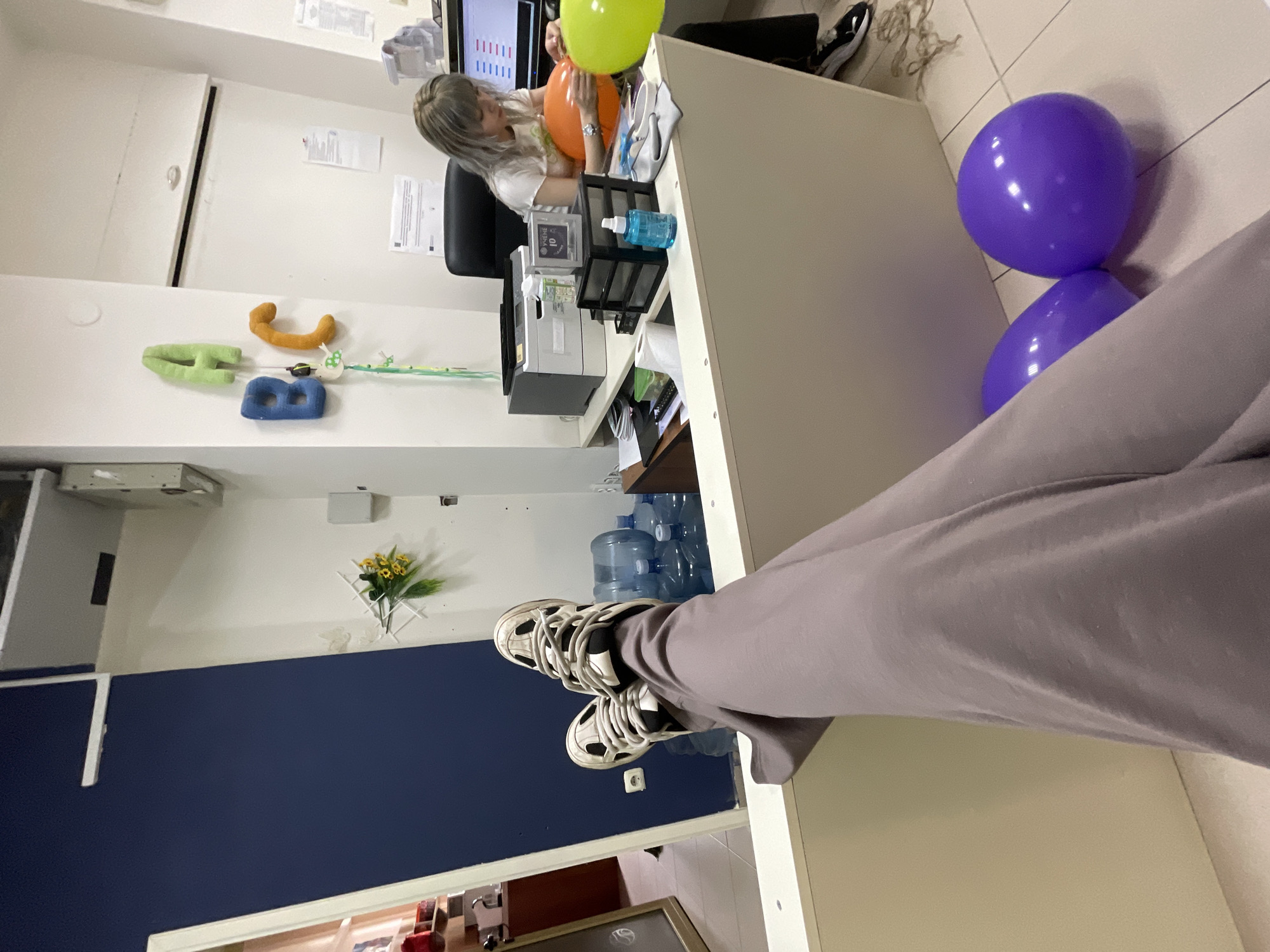 The image displays an interior scene within an office or commercial space in Unknown, Unknown, captured from a first-person perspective. The photographer's legs, dressed in loose grey pants, and feet, wearing white and black athletic sneakers with prominent white laces, are propped up on a light-colored counter in the foreground. Two inflated purple balloons rest on the tiled floor to the right of the photographer's legs.
In the midground, a woman with light-colored hair is seated at a dark desk chair, facing right. She is wearing a white t-shirt and dark pants, and is actively handling an orange balloon on the desk surface. A bright green balloon is partially visible beside it. The desk also contains a grey and white printer, a black multi-drawer organizer, a blue spray bottle, and other office items. A computer monitor displaying a graphic interface is visible behind the seated woman.
The background features a wall section painted dark blue on the left and white on the right. The white wall is decorated with artificial yellow flowers affixed to a white grid, and above them, three padded fabric letters spelling "A", "B", "C" (green, blue, and orange, respectively) are displayed. Several large, clear water cooler bottles are stacked on the floor near the blue wall. The ceiling consists of white panels with integrated lighting.