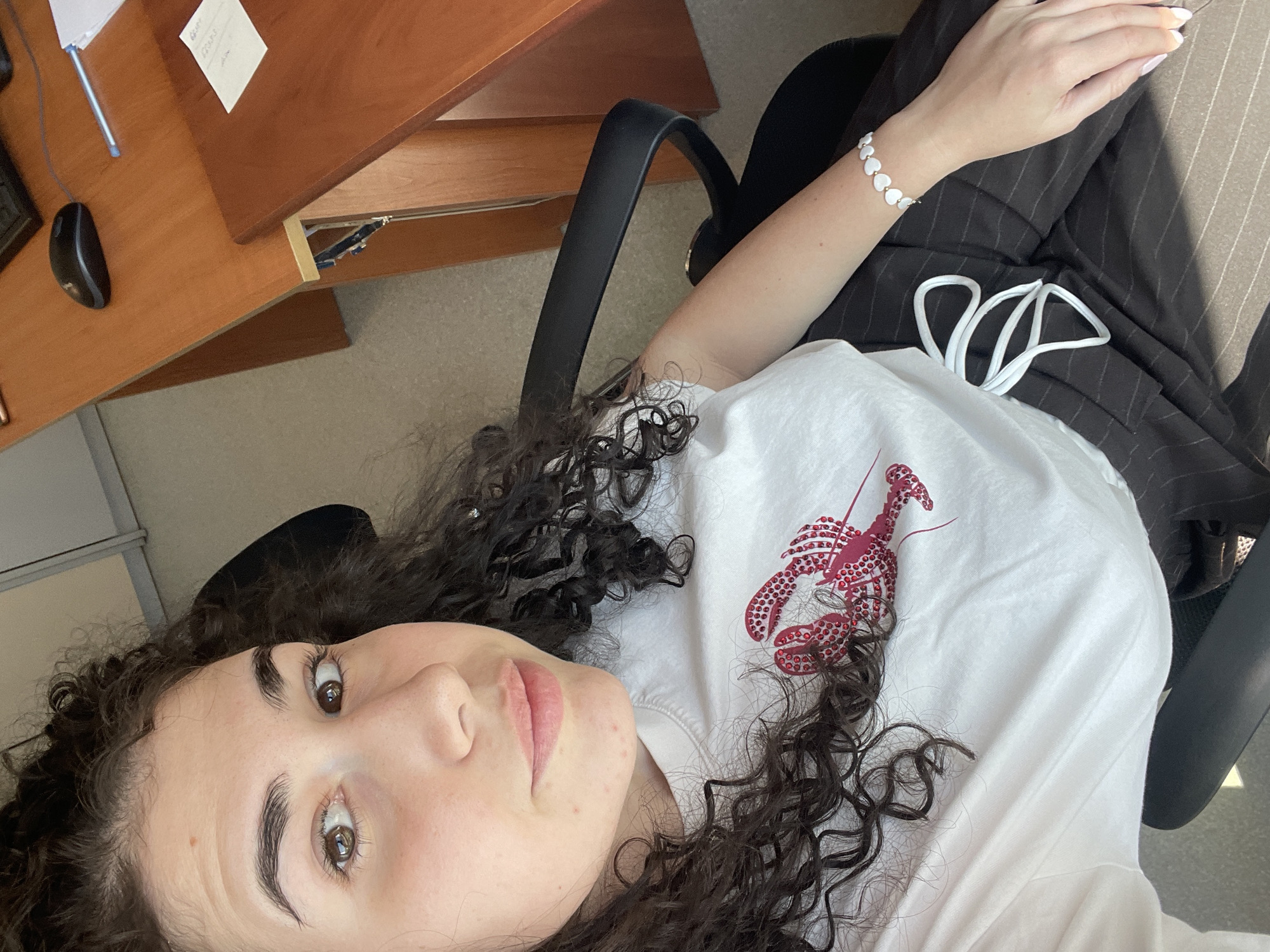 The image captures a young woman in an indoor setting, likely an office or study room, located in Sofia, Bulgaria. The shot is taken from a slightly elevated perspective, with the woman looking directly at the camera while reclining or lying down.
The main subject is a woman with dark, curly hair and brown eyes, visible from the torso upwards. She wears a white short-sleeved t-shirt featuring a prominent red lobster design on the chest, detailed with numerous small, shiny embellishments. A white heart-shaped beaded bracelet adorns her left wrist. She is also dressed in dark pinstriped trousers, visible from the waist down, with a white drawstring detail. Her expression is neutral and direct, gazing upwards at the camera.
In the upper left portion of the frame, a wooden desk is visible. On the desk, there's a black computer mouse, a blue pen, and a few white papers. One paper has some handwritten text, which is largely indecipherable, though fragments like "GIVE" and what appears to be numbers (e.g., "12345") can be faintly discerned. Part of a black office chair is also seen, supporting the woman. The floor beneath her appears to be a light-colored carpet or similar material. The scene is well-lit by natural light, suggesting it is daytime.