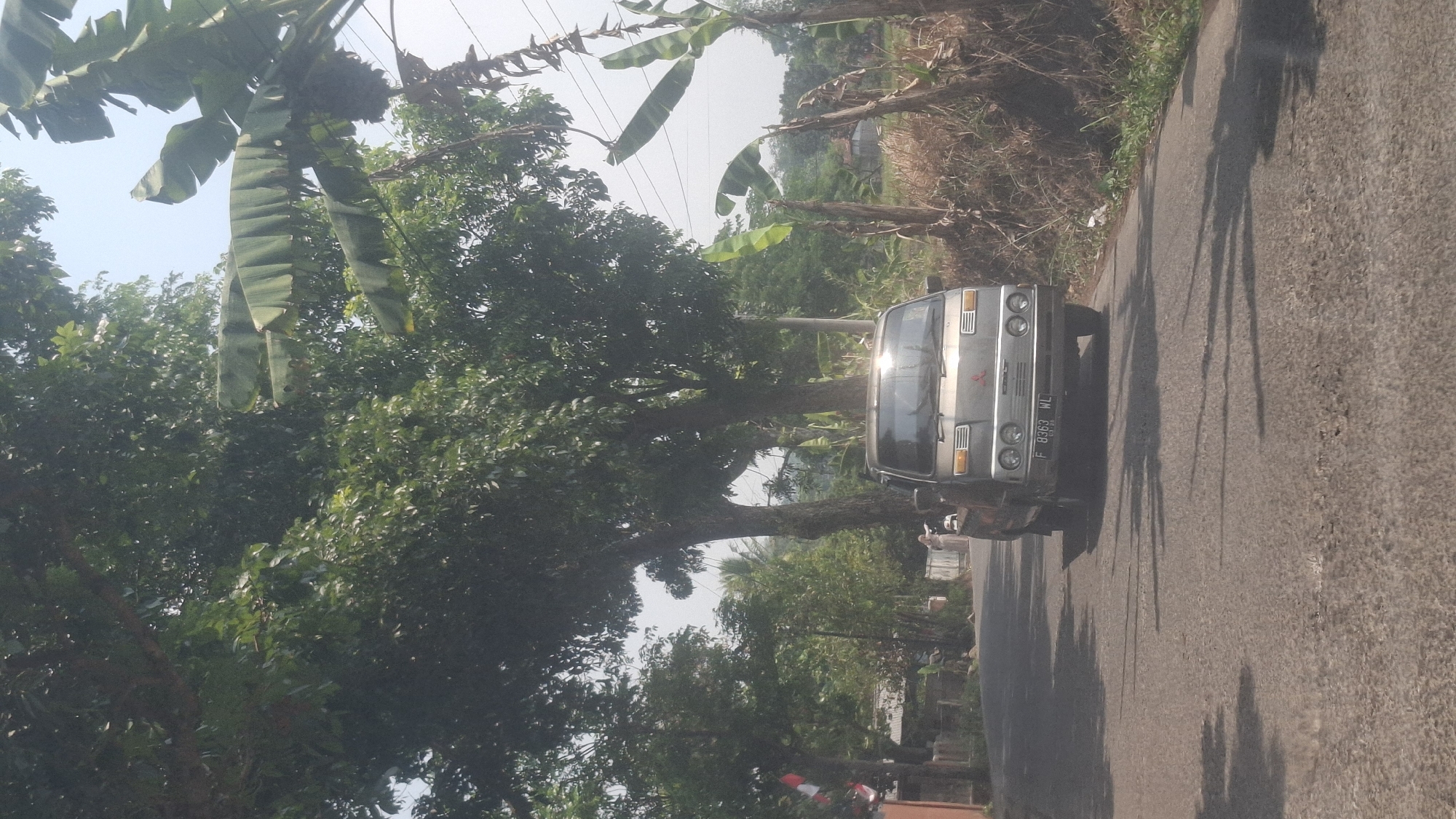 A silver Mitsubishi Colt light truck is driving down a paved road lined with trees and vegetation. The truck's license plate reads "F 8363 WL". In the background, there are houses and more trees, suggesting a rural or semi-rural setting. A person in a light-colored outfit is visible standing near the truck on the left side of the road. The sky is clear and bright, indicating a sunny day. The road surface appears somewhat worn with some cracks and potholes. The scene is likely located in Cibinong Hilir, Indonesia, given the presence of lush greenery and the style of housing visible.
