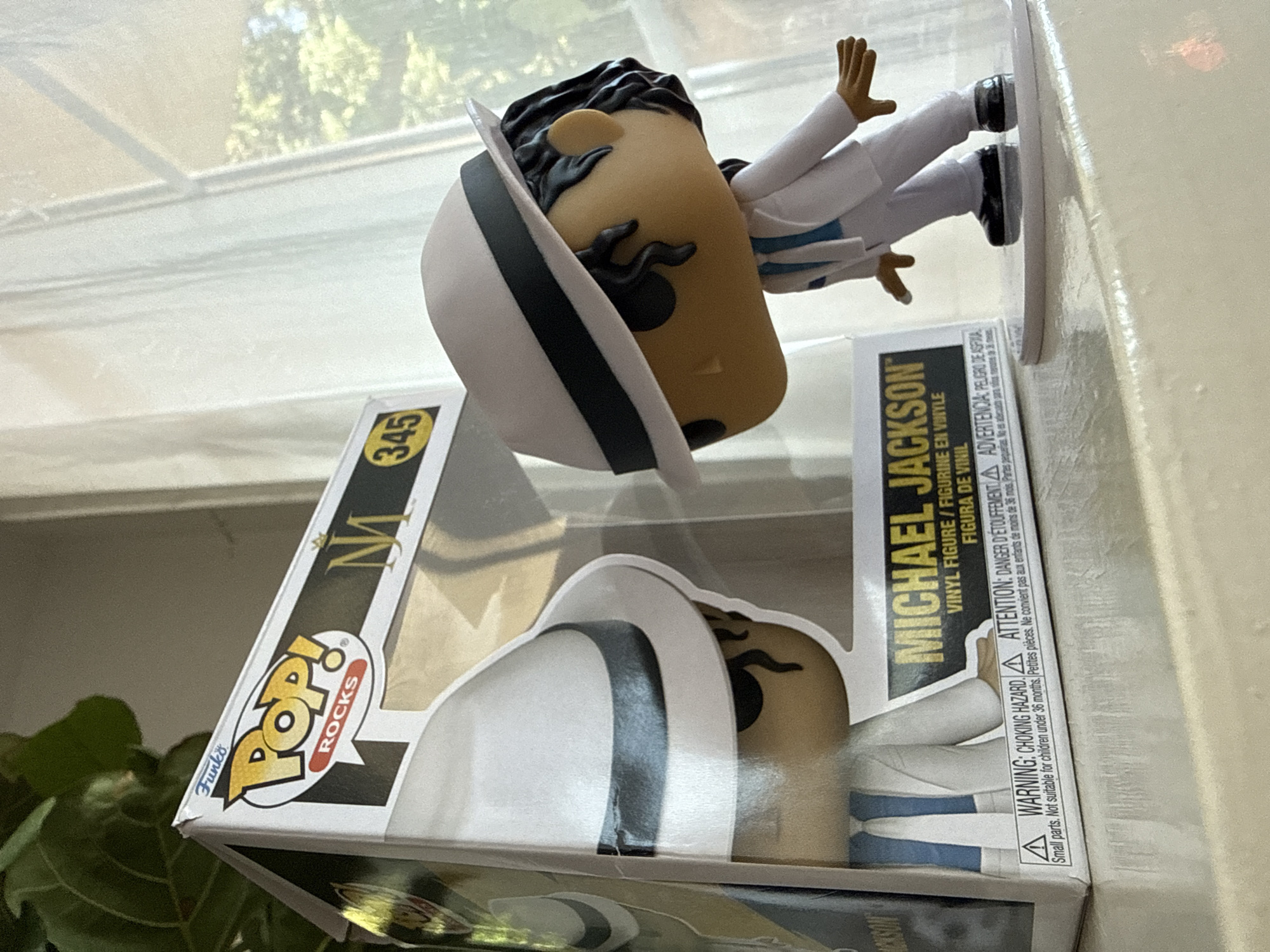 The image shows a Funko Pop! vinyl figure of Michael Jackson, a well-known musician, in its packaging. The figure is wearing a white suit and a white fedora with a black band, characteristic of his "Smooth Criminal" era. The packaging is partially visible on the left side of the image and displays the "Pop! Rocks" series logo and the number 345. The figure is standing on a clear display base. The background consists of a white curtain and a window with a view of trees outside, suggesting an indoor setting, possibly a home. The image is taken from a slightly low angle, emphasizing the figure. There is no indication of the city of Bad Sachsa, Germany, within the provided image.