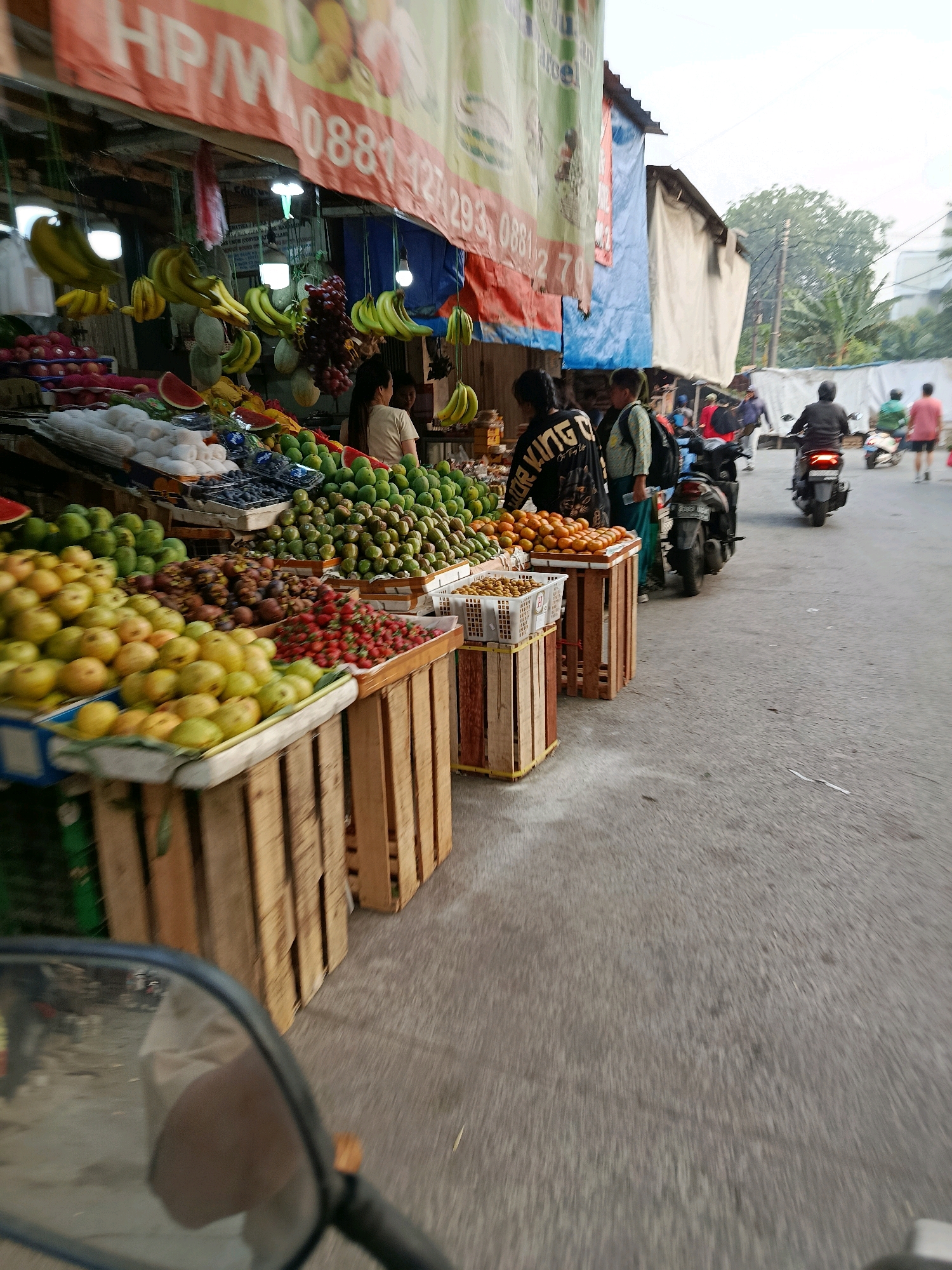 This is a street-level view of a fruit stand in Bekasi, Indonesia, taken from the perspective of a moving vehicle, likely a motorcycle, indicated by the rearview mirror in the foreground. The fruit stand displays an abundant variety of fruits, including bananas, grapes, watermelons, mangoes, guavas, strawberries, and what appear to be mangosteens, arranged in wooden crates and baskets. The stand is adorned with banners, one of which has partial text in Indonesian, including a phone number. Several people are visible at the stand, some appearing to be vendors and others customers. In the background, a street scene unfolds with other pedestrians and several motorcycles, one of which is being ridden. Buildings, some covered with tarps, line the street, and lush green trees are visible in the distance, suggesting an outdoor market setting. The overall impression is a bustling, everyday scene in a market area of Bekasi.