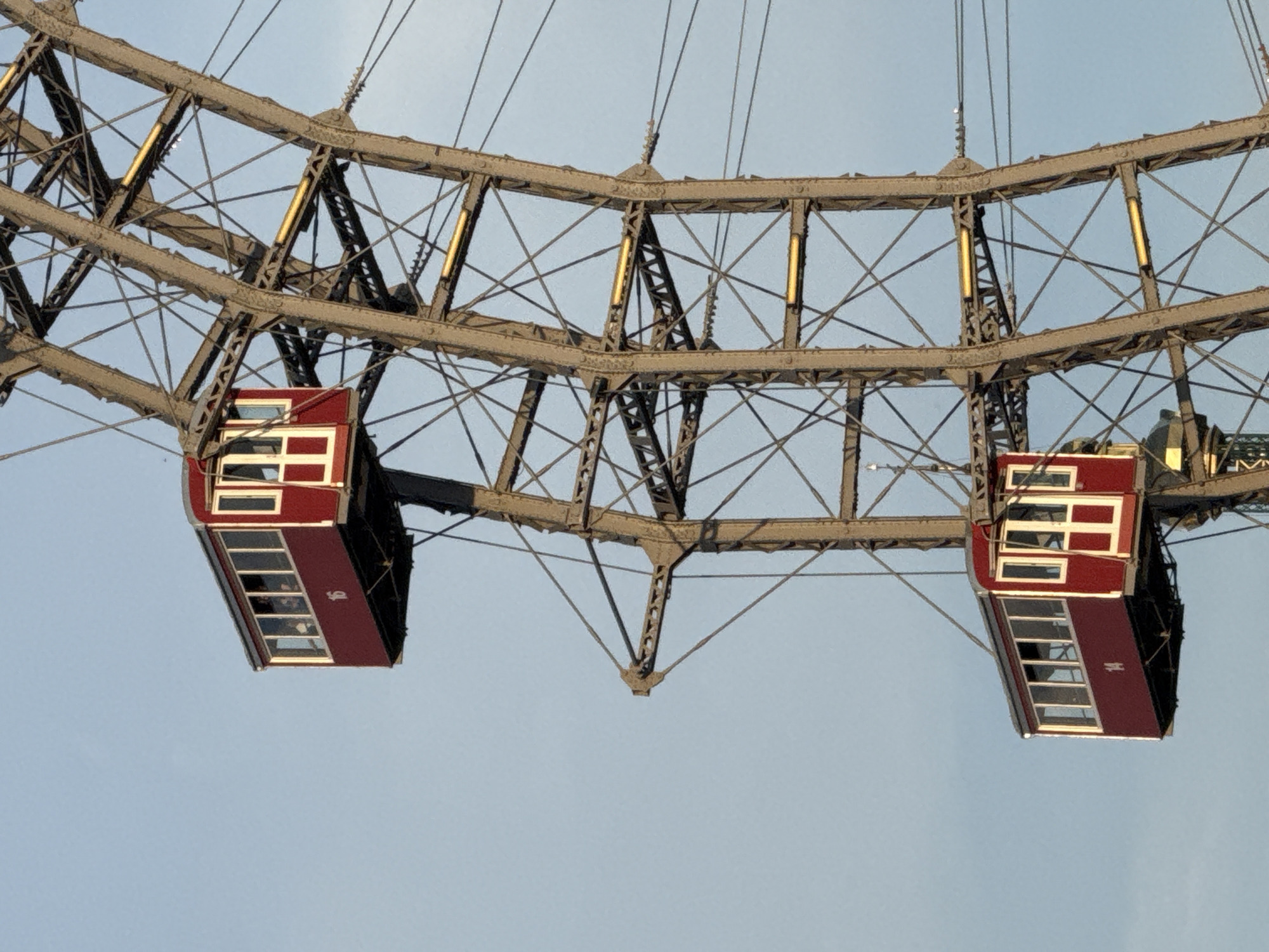 The image shows a close-up of two gondolas on the Wiener Riesenrad, a Ferris wheel located in the Prater amusement park in Vienna, Austria. The gondolas are red with white trim and have large windows. One gondola, numbered "16", is positioned higher than the other, which is numbered "14". Both gondolas are suspended from the metallic structure of the Ferris wheel against a clear blue sky. The metallic structure is made of crisscrossing beams and supports.