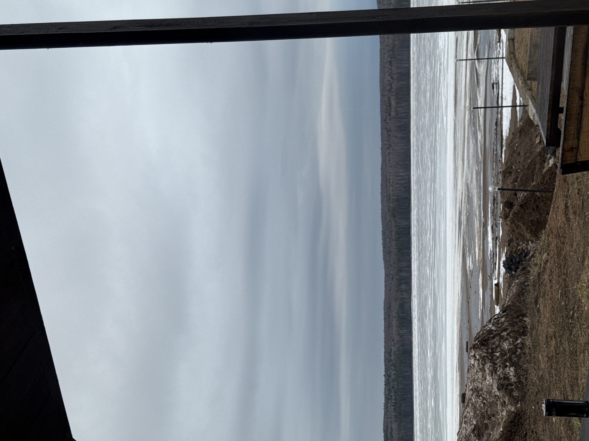 The image depicts a view of the ice-covered Kama River from a structure, potentially a balcony or deck, in Chaikovsky, Russia. The sky is overcast with gray clouds. A dark line of trees is visible on the opposite shore. The foreground includes a section of the deck with its wooden support beams, and a grassy area with patches of dirt and melting snow along the riverbank. Small, evenly spaced decorative light posts stand along the edge of the riverbank. A black cylinder-shaped object is positioned on the ground. There are no people visible in the scene. The dominant impression is of a cold, late-winter day on the river.