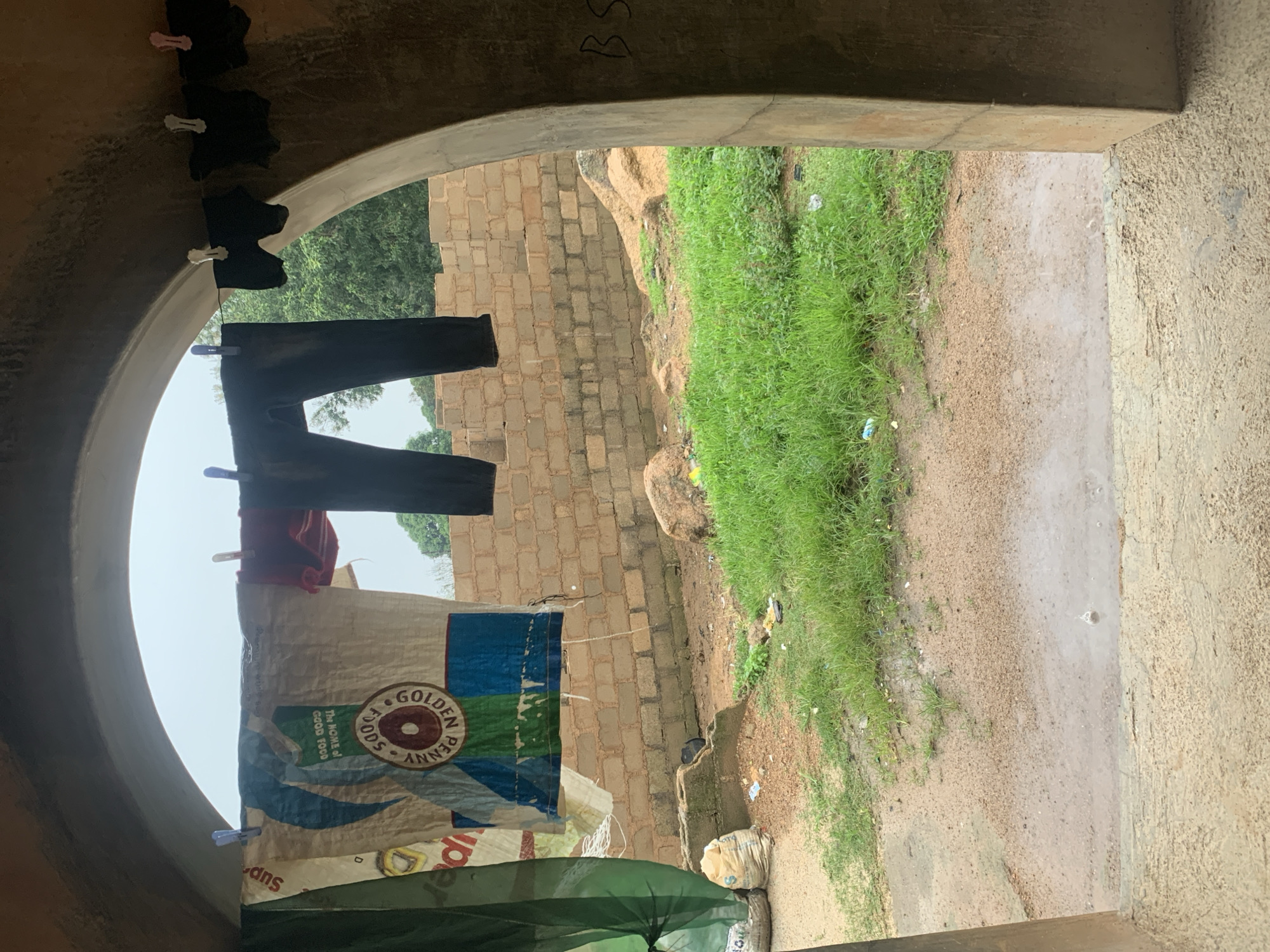 The image depicts an outdoor scene viewed through an arched entryway. In the foreground, a rough, sandy ground transitions to a patch of wet dirt, suggesting recent rain. Lush green grass grows abundantly in the midground, interspersed with some scattered debris. Beyond the grass, a rough brick wall serves as a backdrop, with some unfinished construction elements visible. Large rocks are present among the grass and sand. The sky appears overcast.
Hanging from a clothesline stretched across the entryway are several items of clothing. On the left, a large piece of fabric, possibly a tarpaulin or bag with a "GOLDEN PENNY FOODS" logo, is suspended. Next to it, a pair of dark trousers hangs. To the right, several dark undergarments are also drying. The overall setting appears to be a residential or transitional area, likely within a developing urban environment. The presence of the unfinished brickwork and the humble materials of the hanging clothes suggest a modest dwelling.
The photograph captures a slice of everyday life, focusing on the functional aspect of drying laundry against a backdrop of raw, unrefined construction. The scene is devoid of any people, and the focus is on the objects and the immediate environment. The light is diffused, consistent with an overcast day. The image offers a glimpse into the visual texture and atmosphere of a specific, likely non-descript, location within the city of Jos, Nigeria, characterized by its utilitarian structures and natural elements.