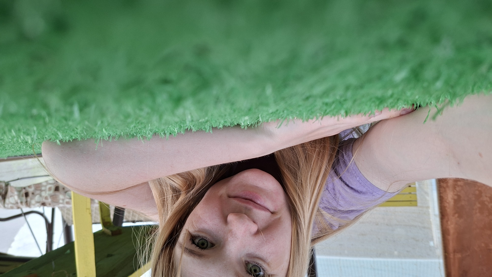 A young Caucasian female with long blonde hair and green eyes is lying down on artificial grass, looking directly at the camera. She is wearing a purple t-shirt and resting her head on her arms. The background shows outdoor elements including a brown wall on the left and a yellow structure with some draped fabric on the right, suggesting an outdoor recreational area possibly in Большой Букор, Russia. The perspective is low, emphasizing the texture of the artificial grass.