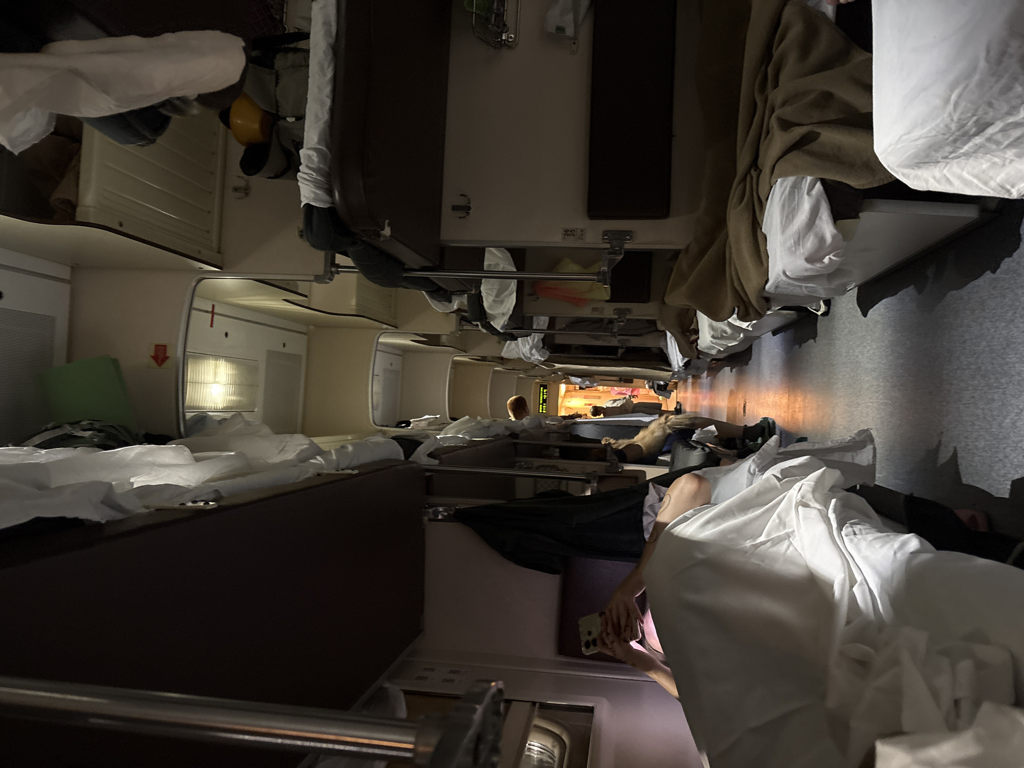 The image shows the interior of a compartment on a train, likely a sleeper car, with bunk beds lining the walls. In the foreground, a person is lying on a lower bunk, holding a smartphone. The compartment is dimly lit, with some overhead lights and light coming from further down the train car. Several beds are made up with white linens and brown blankets. Clothes are hanging on hooks and draped over bunks. The overall environment suggests a nighttime journey. The location is identified as Inza, Russia, which is a city in Penza Oblast, Russia, known for its railway junction.