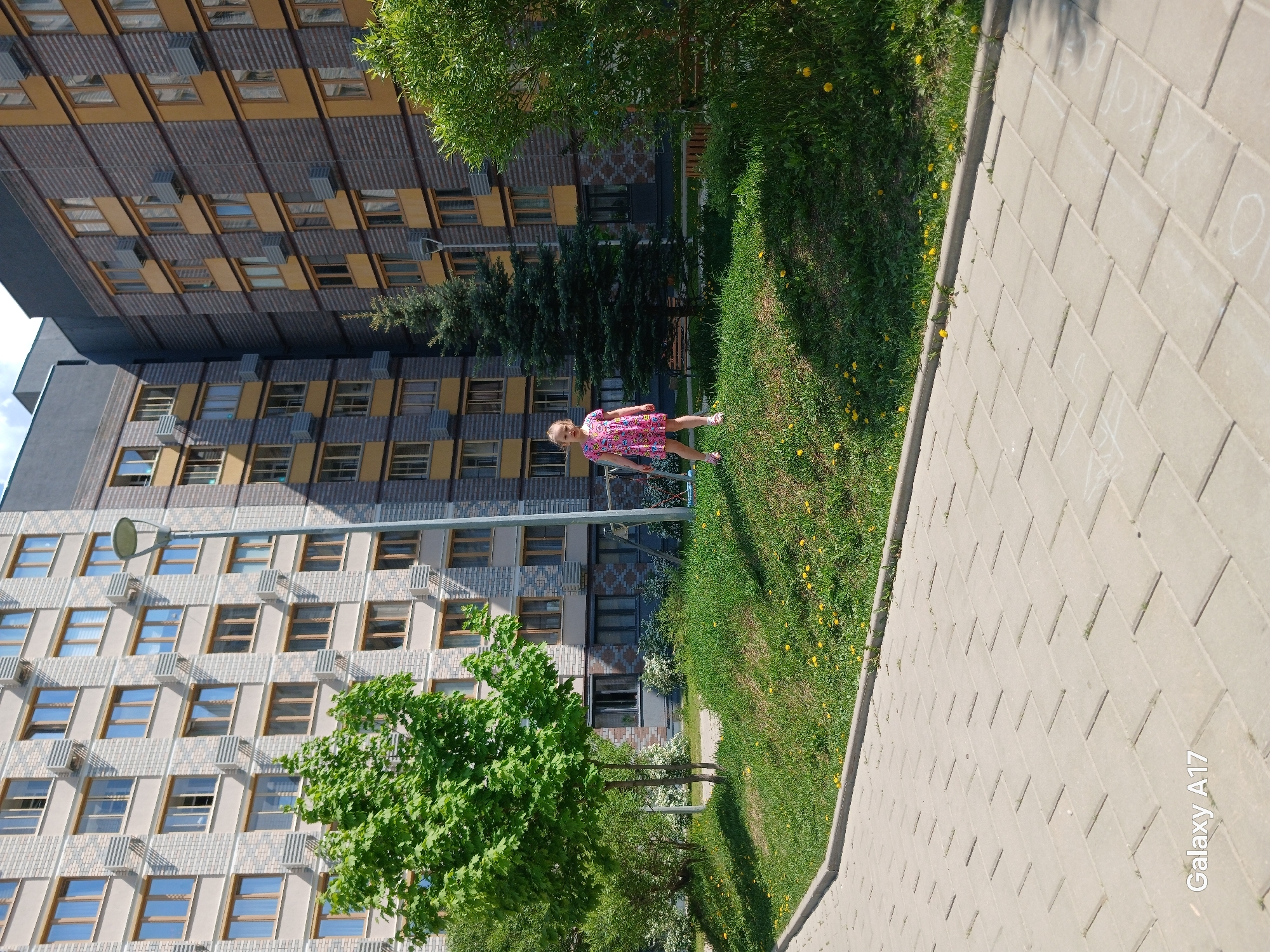The image shows a girl in a pink dress walking on a grass lawn next to a sidewalk, with a multi-story building in the background. The building has a light gray facade with rows of rectangular windows framed in brown. There is some greenery, including trees and dandelions, visible in the lawn. The photograph was taken in Yekaterinburg, Russia.
