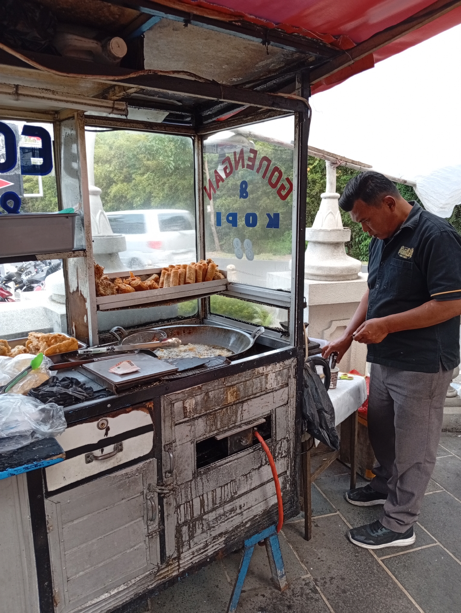 An Indonesian man stands by a street food cart in Cimacan, Indonesia. The cart is equipped with a deep fryer and displays various fried foods, including spring rolls and chicken. The man is wearing a black collared shirt and grey pants, and he is holding a small item in his right hand, appearing to interact with it. The background shows lush green trees and a white vehicle. The scene suggests an active food vendor operating in a public space within the city.