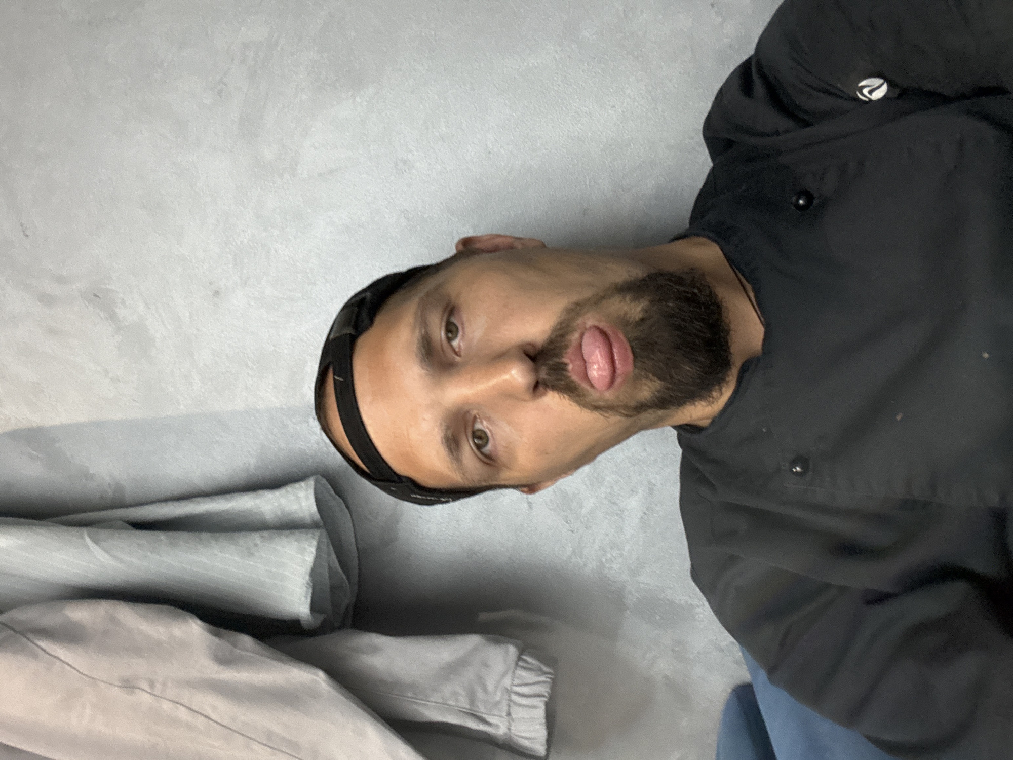 A man with a goatee and a black baseball cap worn backward is sticking out his tongue at the camera. He is wearing a black chef's coat. The man appears to be sitting down, with a blue couch visible in the foreground. Behind him, a grey, textured wall is visible, with a light-colored jacket hanging on the left side of the frame. The image appears to be taken indoors. The location is not specified as Sochi, Russia, nor any other specific city.