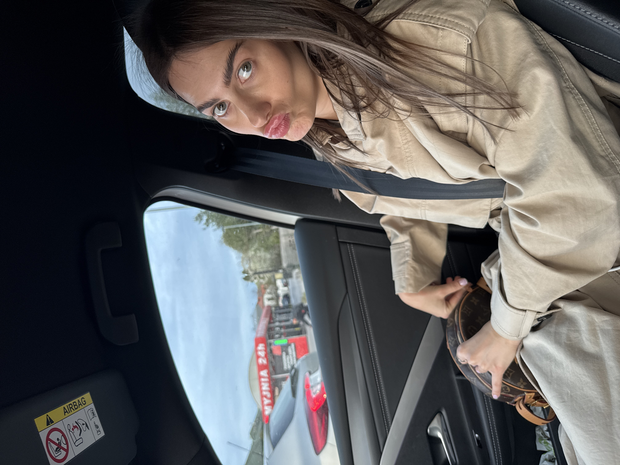 A woman with long brown hair sits in the passenger seat of a car. She wears a tan trench coat and a seatbelt, and she has a brown and tan patterned handbag in her lap. She pouts her lips toward the camera. Outside the car window, traffic is visible, along with a sign in Polish that reads "WYJMNIA 24h," indicating a vehicle recovery service operating 24 hours a day. The setting is Warsaw, Poland.