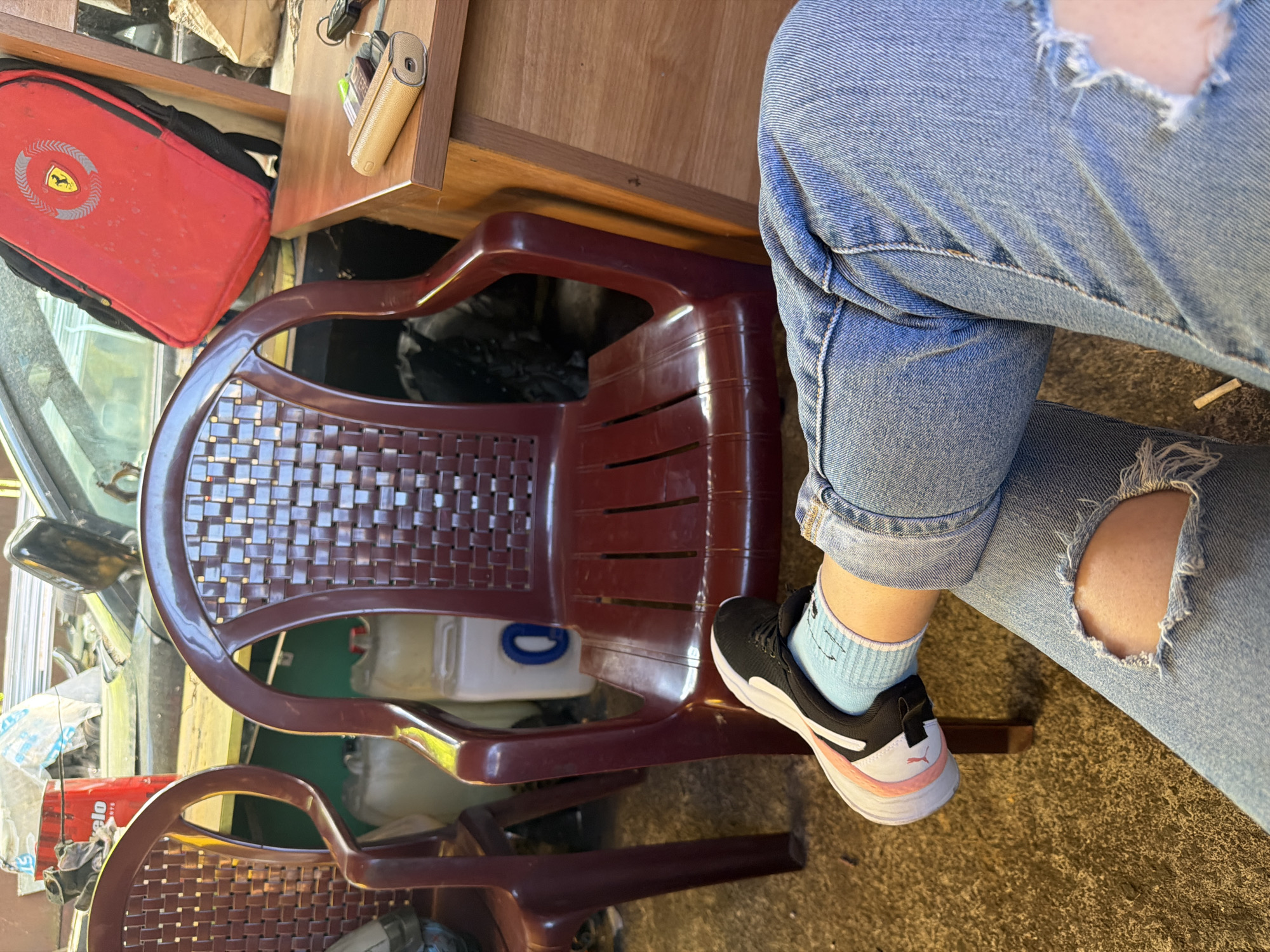 The image depicts a person's legs and feet, wearing ripped blue jeans and light blue socks with black and white sneakers, seated in front of a dark red plastic chair. The chair has a woven-patterned back and seat. Behind the chair, a red backpack with a Ferrari logo is visible, leaning against a wooden desk. Various objects, including what appear to be plastic containers and a metal can, are scattered in the background, suggesting an outdoor or semi-outdoor storage area. The flooring is a coarse, textured material. The scene does not provide specific visual cues to pinpoint the location in Burgas, Bulgaria, but the presence of plastic chairs and containers could be indicative of an outdoor seating area or a storage space commonly found in such a city.
