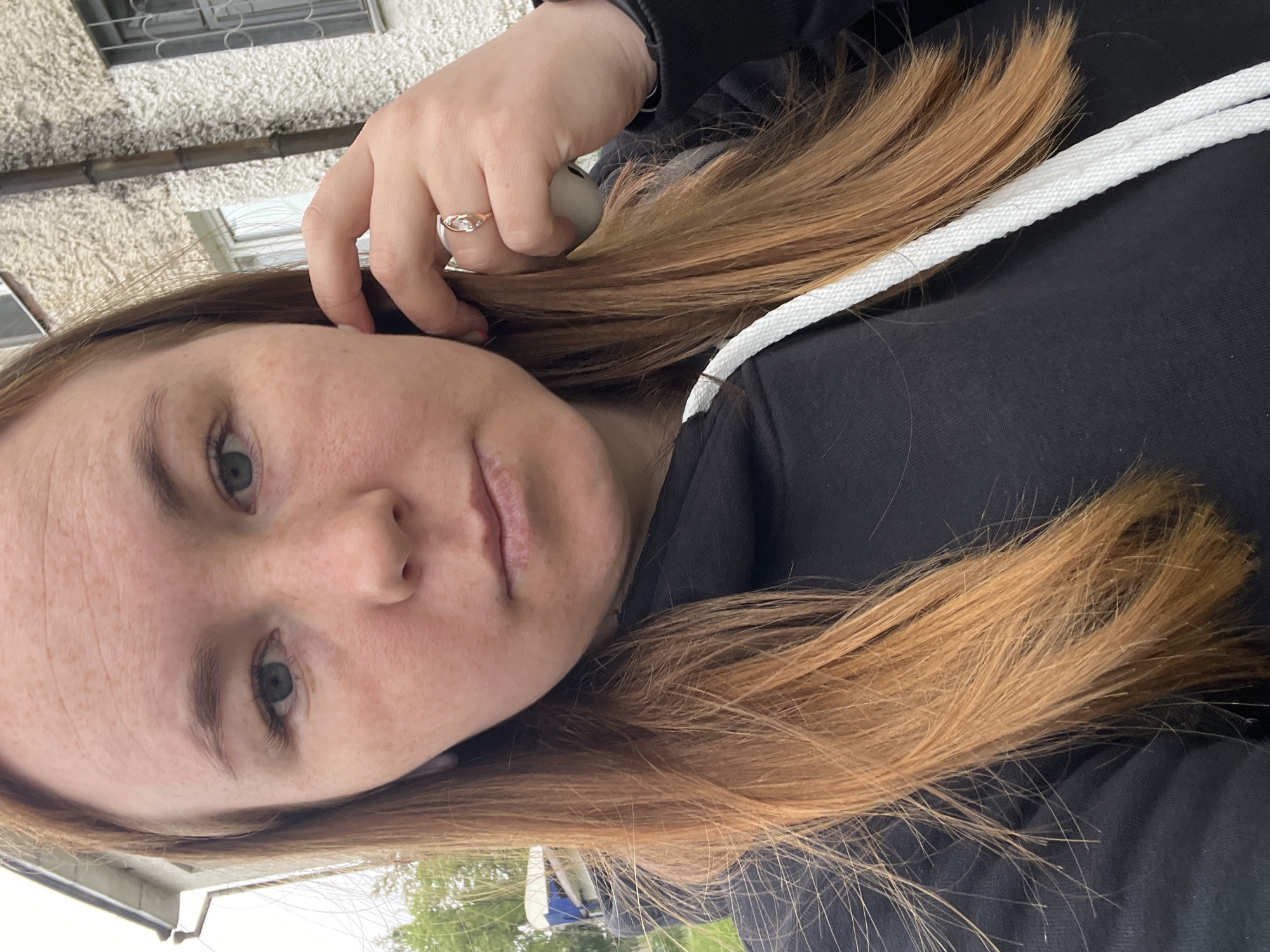 This close-up selfie, taken in Chaikovsky, Russia, features a person, likely female, with fair skin and noticeable freckles, green/hazel eyes, and long, reddish-brown hair. The subject wears a black hooded sweatshirt with a prominent white drawstring visible across the chest. Her expression is neutral.
Her right hand is raised towards her ear, with fingers gently touching her temple or hair. A golden ring with a small, clear or white stone is visible on her ring finger. Partially obscured by her hand, a white, rounded object, possibly an earbud, can be seen near her ear.
The background is an outdoor setting, mostly out of focus. Behind the subject's head, a light-colored, textured building wall, possibly stucco, is visible. Above this wall, a window with metal bars or a grille can be discerned. Further in the background, there are hints of green foliage from trees and other indistinct building structures, all under natural daylight, suggesting it is daytime. No text is visible in the image.