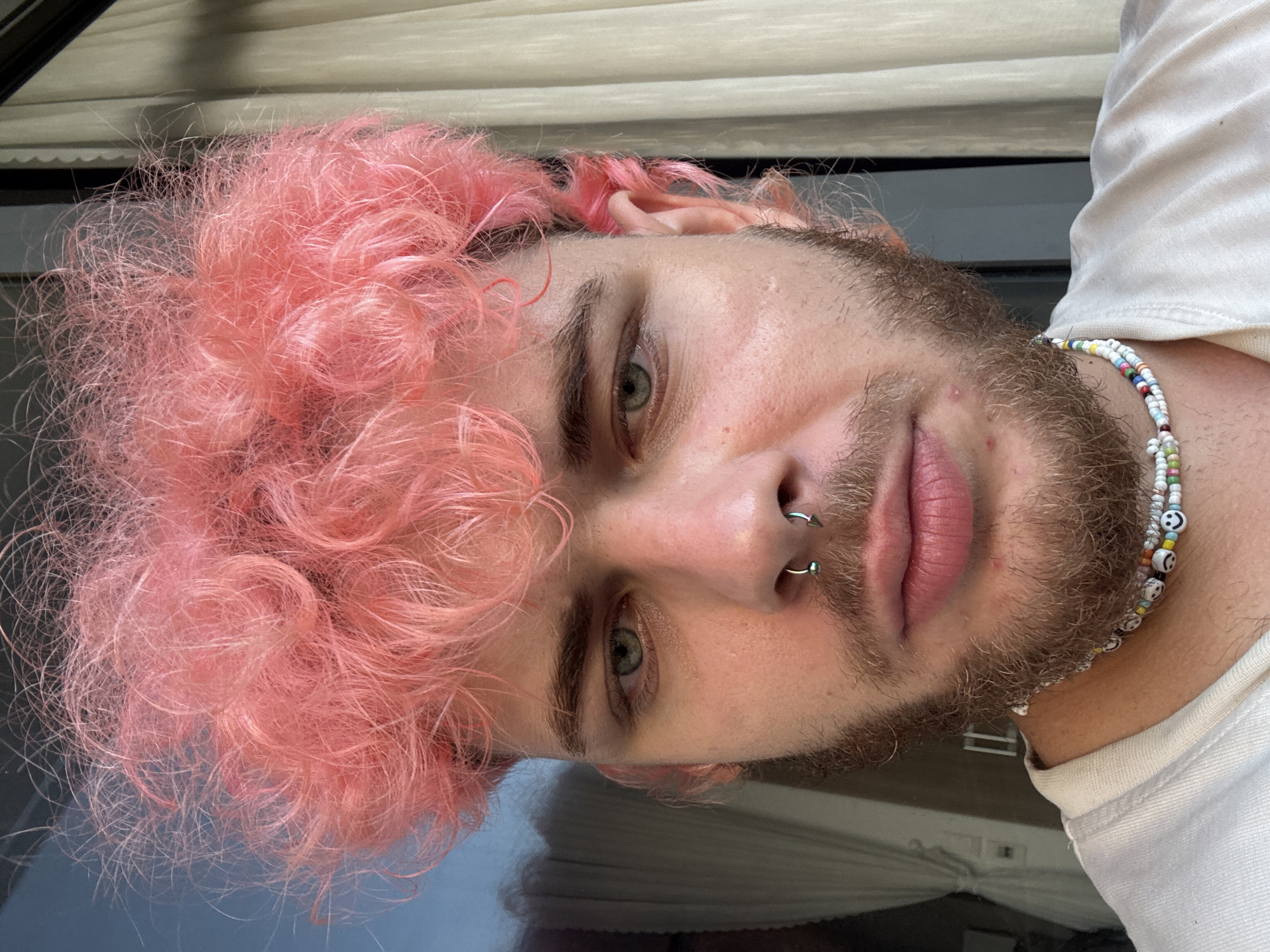 A close-up, indoor portrait shows a person with light skin, hazel eyes, and pink curly hair. The person has a short, brown beard and a double nose ring in their left nostril. They are wearing a white t-shirt and a beaded necklace with smiley face charms. The background appears to be an interior space with window blinds visible. The image provides no information to suggest a location in Đà Nẵng, Vietnam.