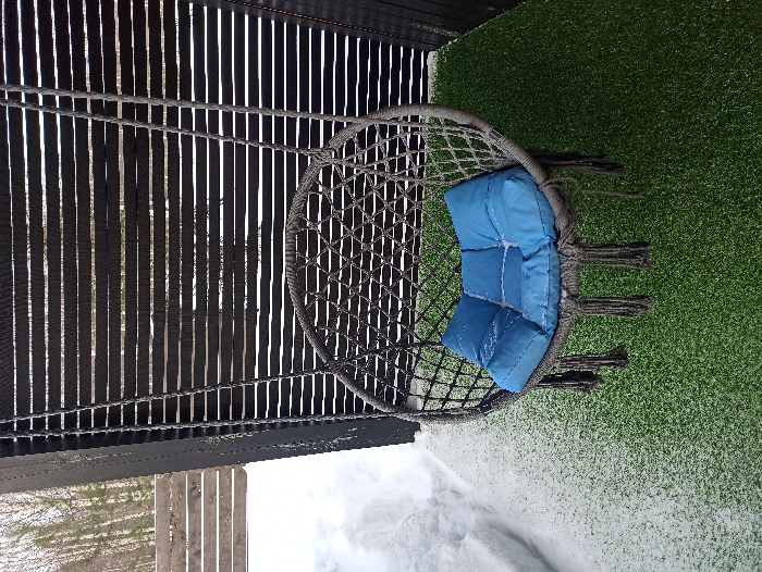 The image displays an outdoor setting in Nizhneye Zadolgoye, Russia. A grey, circular macrame-style hanging chair, adorned with tassels, is centrally positioned. Inside the chair, a segmented blue cushion exhibits light dusting of white snow or frost. The chair is suspended from an unseen overhead structure, positioned in front of a dark, horizontally slatted partition. The ground beneath the chair is covered with artificial green turf, which is sparsely coated with white snow or frost. To the immediate left of the turf, a significant accumulation of white natural snow is present. In the background, beyond the slatted partition and the snow, a sparse stand of bare trees is visible. No individuals or animals are present, and no specific actions or interactions are depicted.