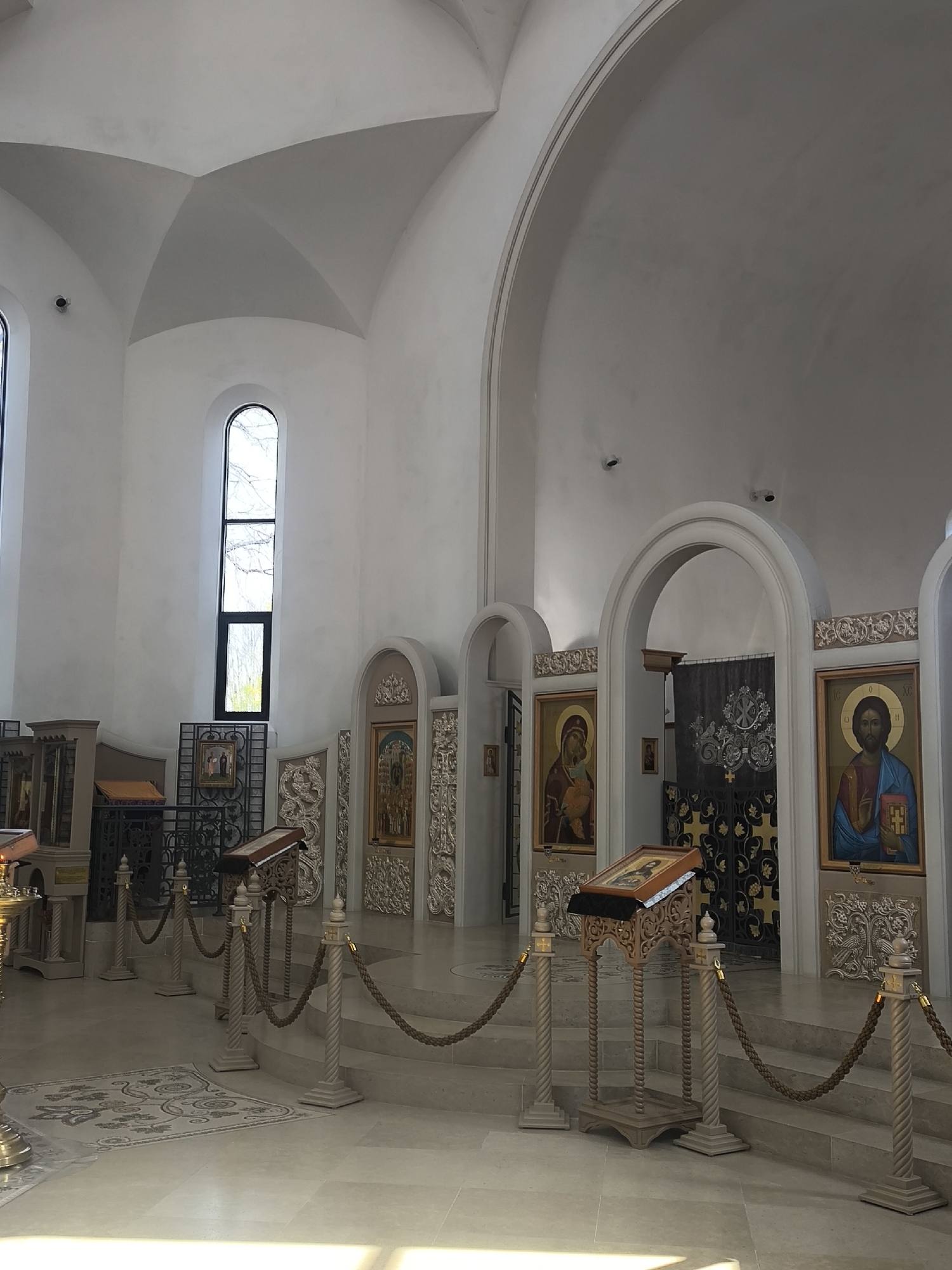 The image depicts the interior of the All Saints Church in Strasbourg, France. It showcases a predominantly white interior design with vaulted ceilings and arched entrances. Religious iconography in the form of paintings are displayed within ornate frames along the walls. Lecterns are placed in the foreground, and stanchions with ropes are used to guide the flow of foot traffic. A tall, arched window provides natural light. The scene appears to be a quiet moment inside the church.