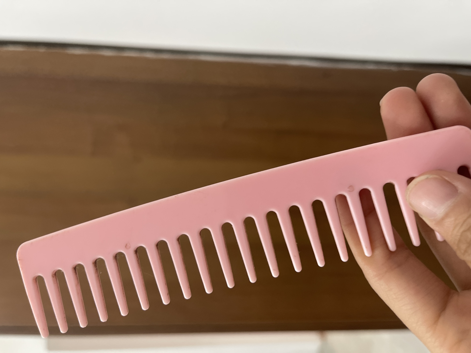 A close-up, slightly angled shot shows a person's hand holding a bright pink plastic comb. The comb is positioned vertically, with its teeth pointing upwards and to the left. The hand is visible at the bottom of the frame, with fingers gently gripping the handle of the comb. The background consists of a wooden door or panel with a warm, brown hue, and a sliver of a white wall to the right. There is no indication of the city of Bishkek, Kyrgyzstan in this image.