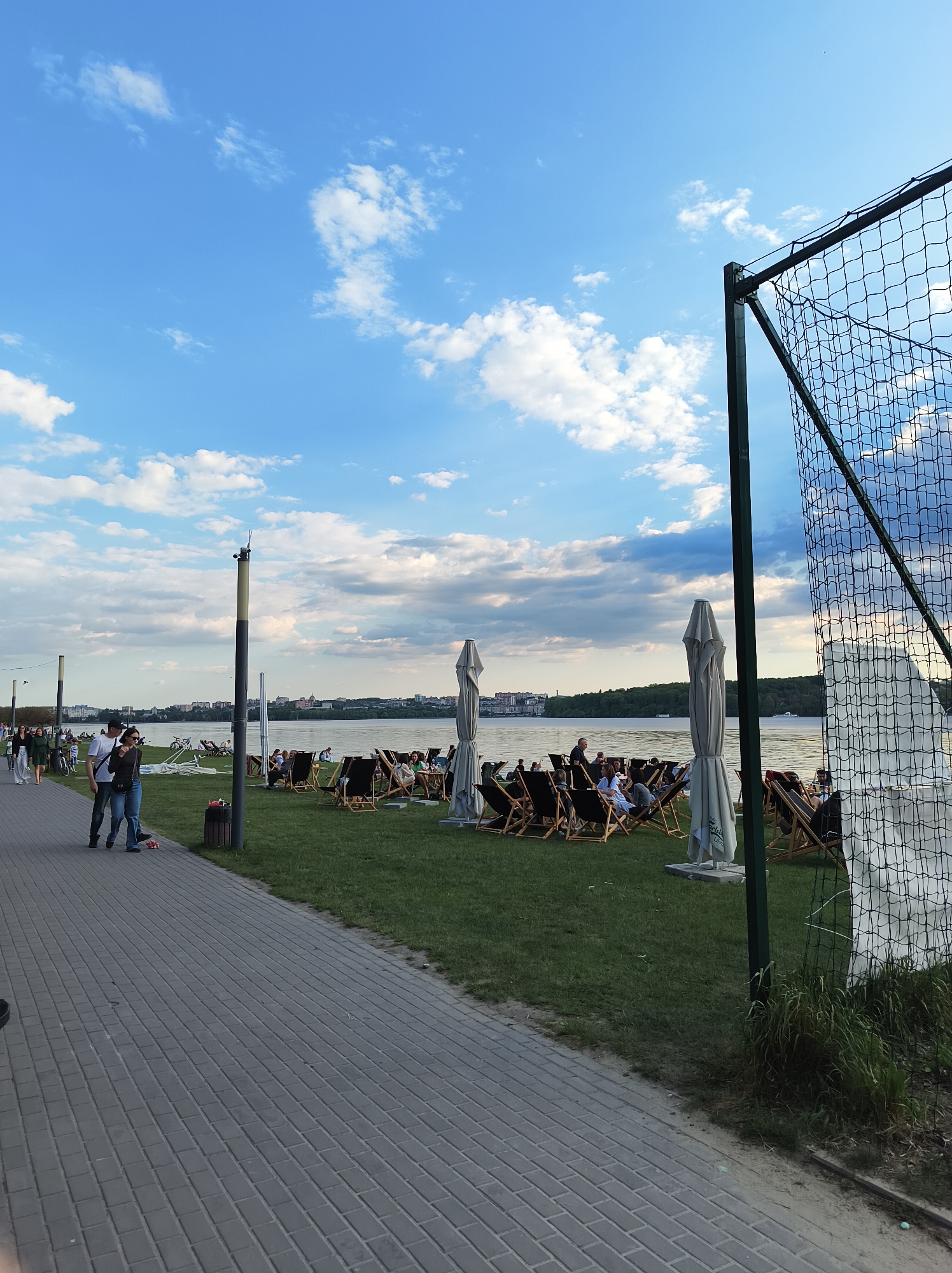 The image captures a waterfront scene in Ternopil, Ukraine. A paved walkway runs along the left, with people strolling along it. Adjacent to the walkway is a grassy area populated with numerous wooden lounge chairs, many of which are occupied by individuals relaxing and facing the water. Several closed white umbrellas are interspersed among the chairs, presumably for shade. The water's edge is visible in the midground, with the cityscape of Ternopil visible in the distance across the water. A tall chain-link fence stands on the right side of the frame, partially obscuring the view. The sky is a mix of blue and white clouds, suggesting a pleasant weather.