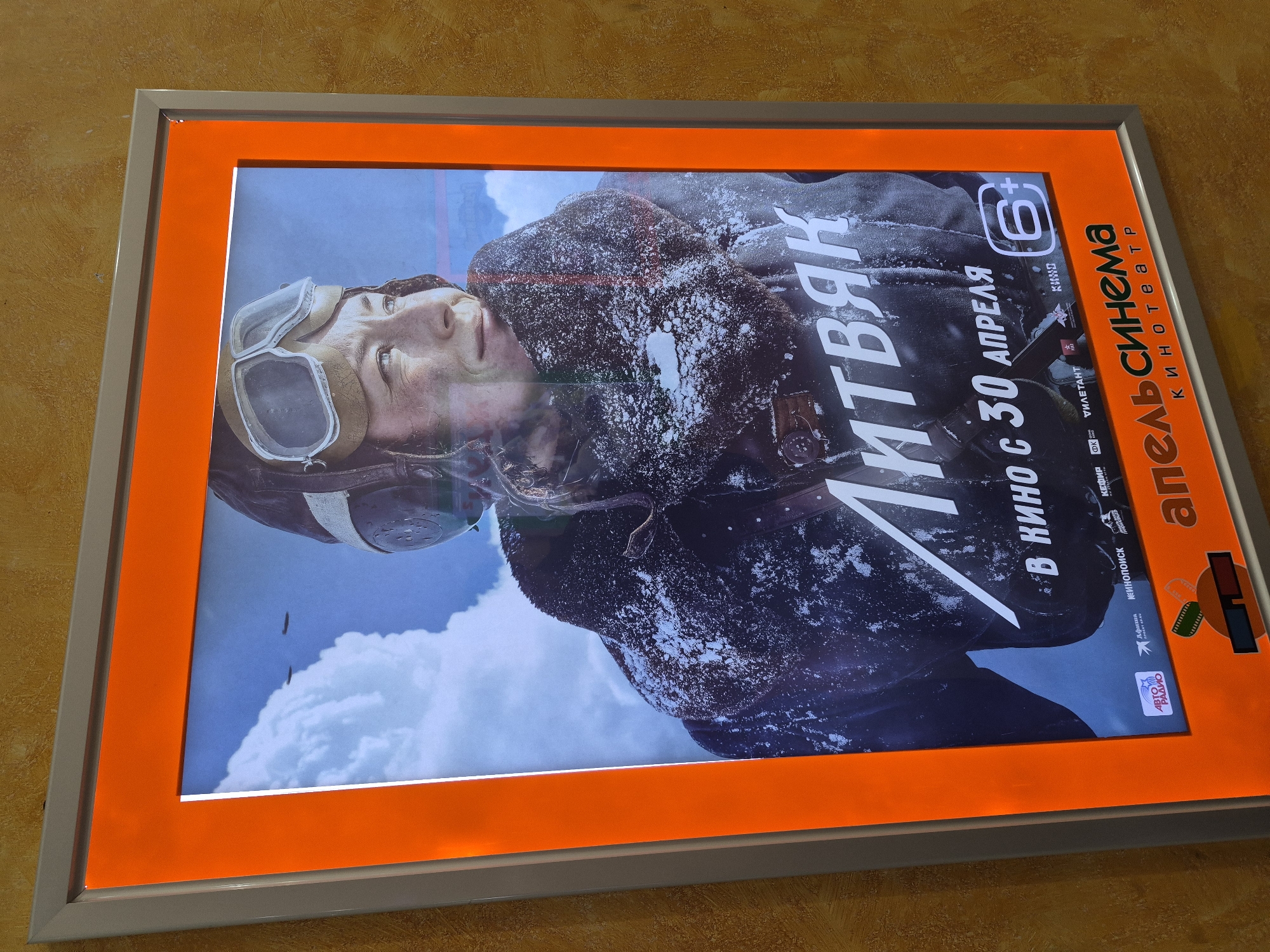 A close-up shot depicts a movie poster of the film "Litvyak" within the "Apelsin Cinema" located in Tobolsk, Russia. The poster features a pilot wearing goggles, with snow apparent on the jacket and helmet, along with the film title in Cyrillic. The text on the poster states the movie will be showing from April 30th, along with a "6+" age rating. Various logos, including "Kinopoisk," "Afisha," "Auto Radio," and others, are also visible. The poster is framed with a vivid orange border and an outer, silver frame.