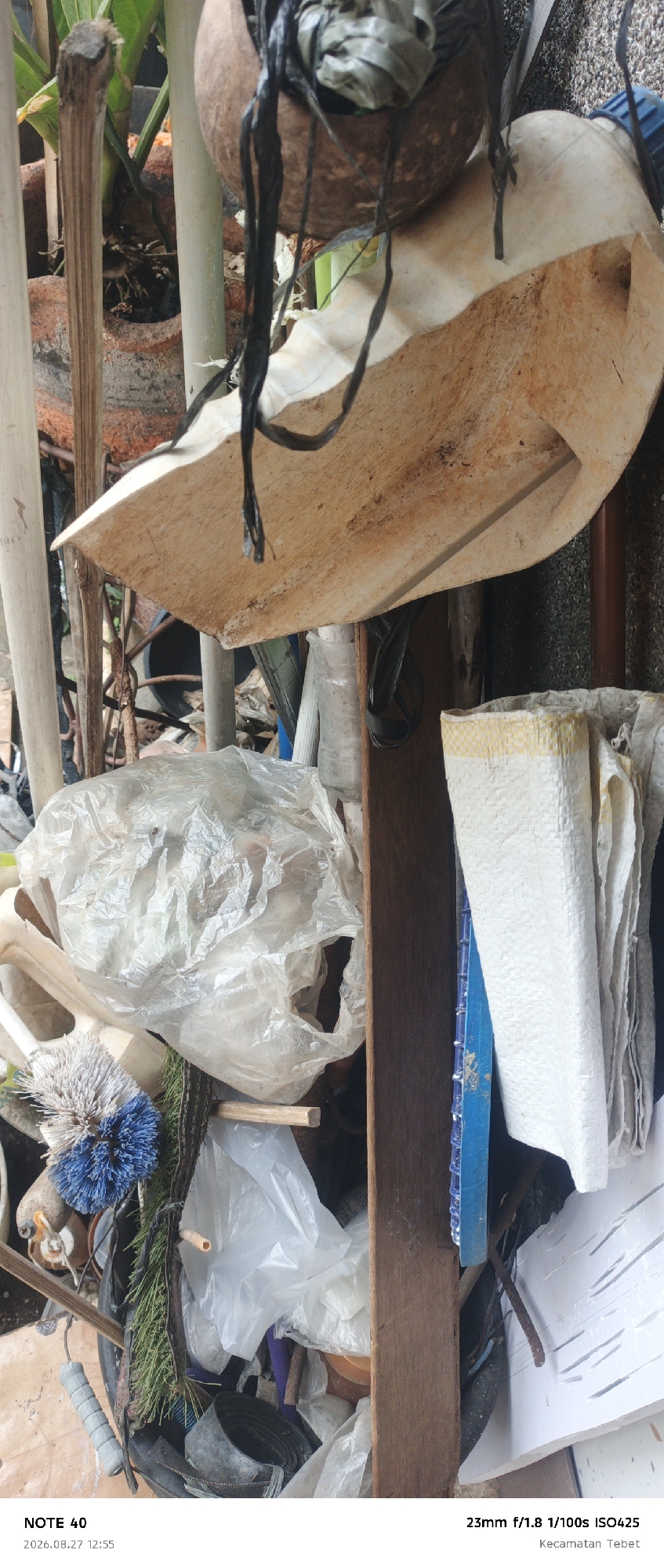 A close-up, vertical, outdoor shot shows a pile of assorted debris. In the foreground, a crumpled translucent plastic bag is prominent. To its left, a blue and white brush head is visible. To the right of the bag, a textured white woven bag and a piece of blue plastic grating are stacked. A vertical wooden plank runs down the center of the image, separating the plastic bag from the woven bag and blue grating. In the upper portion of the image, a large, irregularly shaped object made of a rough, beige material, possibly plastic or composite, is suspended by black cords. A brown, spherical object, possibly a coconut shell, is also suspended nearby. The background is blurred, showing vertical poles and foliage. The image is tagged as being taken in Kecamatan Tebet, South Jakarta, Indonesia.