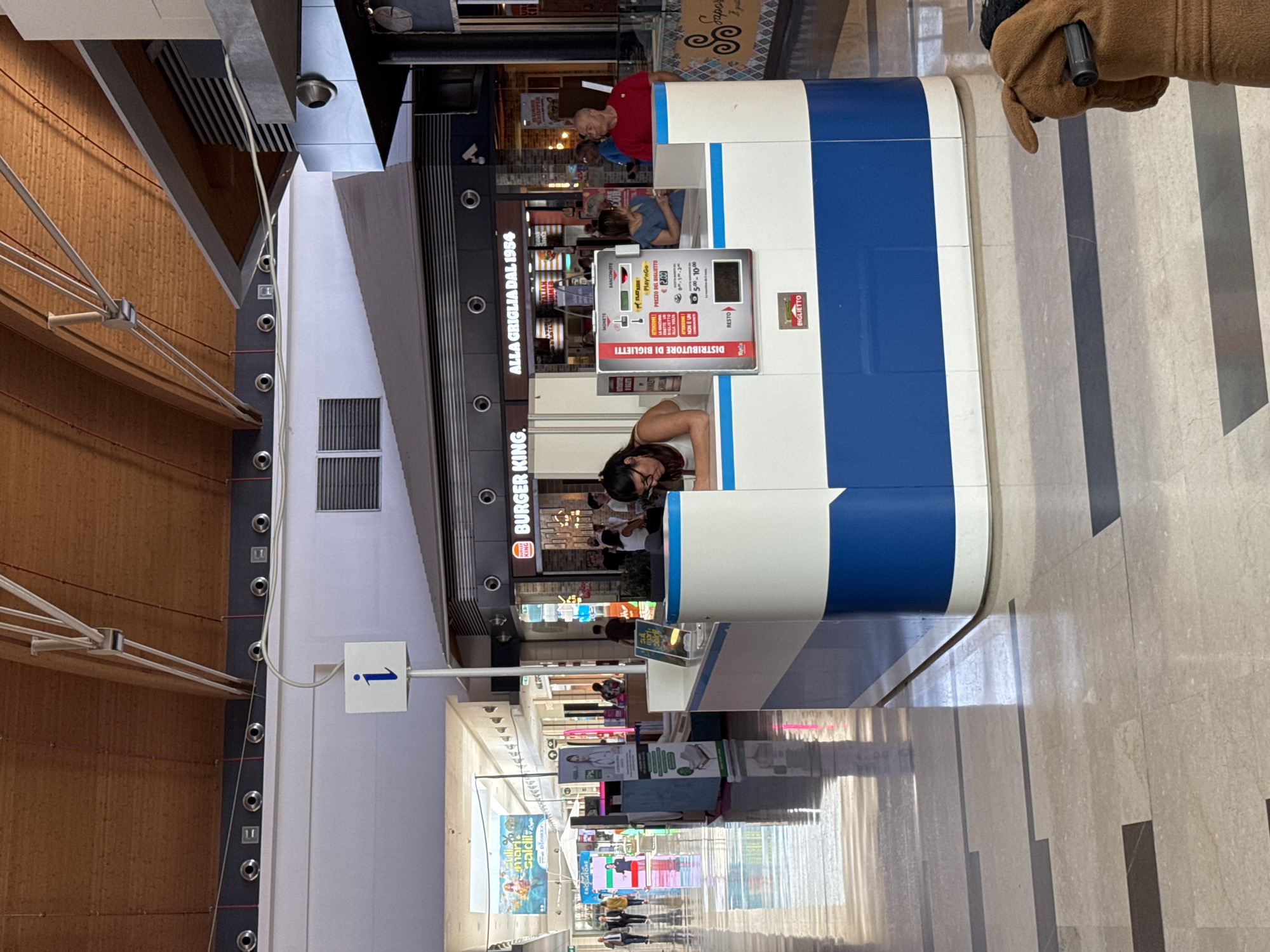 In a brightly lit indoor shopping mall, a ticket vending machine is prominently displayed at a service counter in the foreground. The machine, featuring Italian text and a graphical display of currency options, is situated within a modern blue and white kiosk. A woman with dark hair, wearing glasses and a white top, is leaning over the counter, appearing to interact with the machine. Behind her, the Burger King restaurant is visible with its distinctive signage. Other shoppers are seen in the background, moving through the mall's wide, tiled walkways. The environment is characterized by a high ceiling with exposed wooden beams and industrial lighting. The mall is located in the city of Carini, Italy.