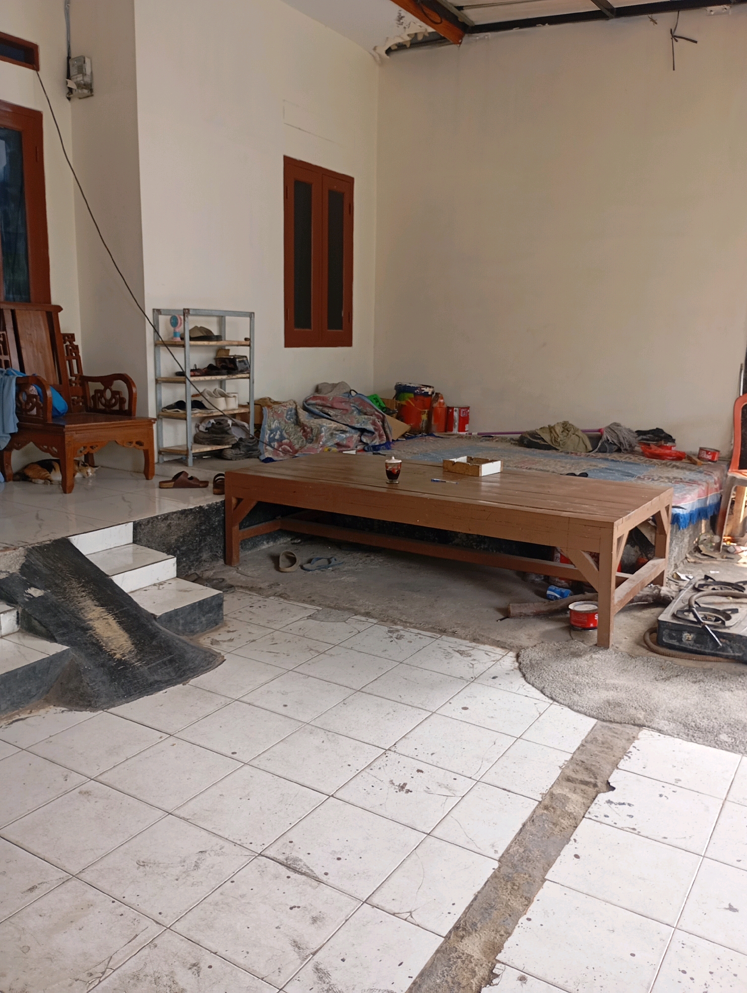 The image depicts an outdoor area of a dwelling in Babelan, Indonesia. The scene is characterized by a low-lying structure with an open-air concept. In the foreground, white tiled flooring is visible, showing signs of wear and tear with scattered dirt and a noticeable trough running through the tiles. To the left, a set of concrete stairs with a black tarp leading down is present. A wooden chair with intricate carvings and a cat resting underneath it are also situated on a slightly elevated tiled area to the left.
Further into the scene, a low, wooden table with two glasses of drinks and a box is placed on a patterned rug. Behind the table, a mattress covered with blankets and assorted items suggests a sleeping or lounging area. To the left of the mattress, a multi-tiered metal shelf holds various items, including shoes and a small fan.
The walls of the structure are painted a light color, and wooden-framed windows are visible. Several cans, possibly paint, are scattered around the sleeping area. A portable gas stove is positioned on the right side of the image, indicating ongoing or recent activity. The overall impression is one of a functional, lived-in space, possibly undergoing some renovation or in a state of disarray.
