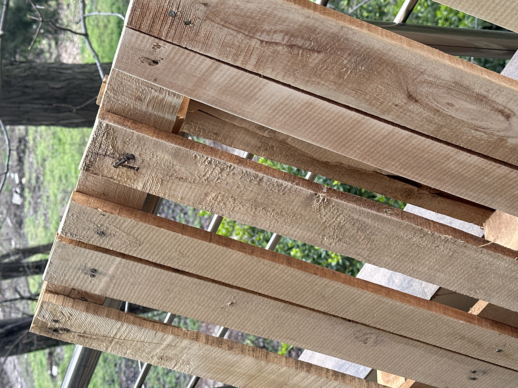 A wooden pallet is shown outdoors, possibly elevated slightly due to the visible metal framework beneath it. The pallet's wood is rough and weathered, with some nails visible. Through the gaps in the pallet, greenery and what appears to be a tree trunk are visible. The image suggests an outdoor setting, potentially a garden or construction area. The location is presumed to be Sofia, Bulgaria, though nothing in the image directly confirms this.