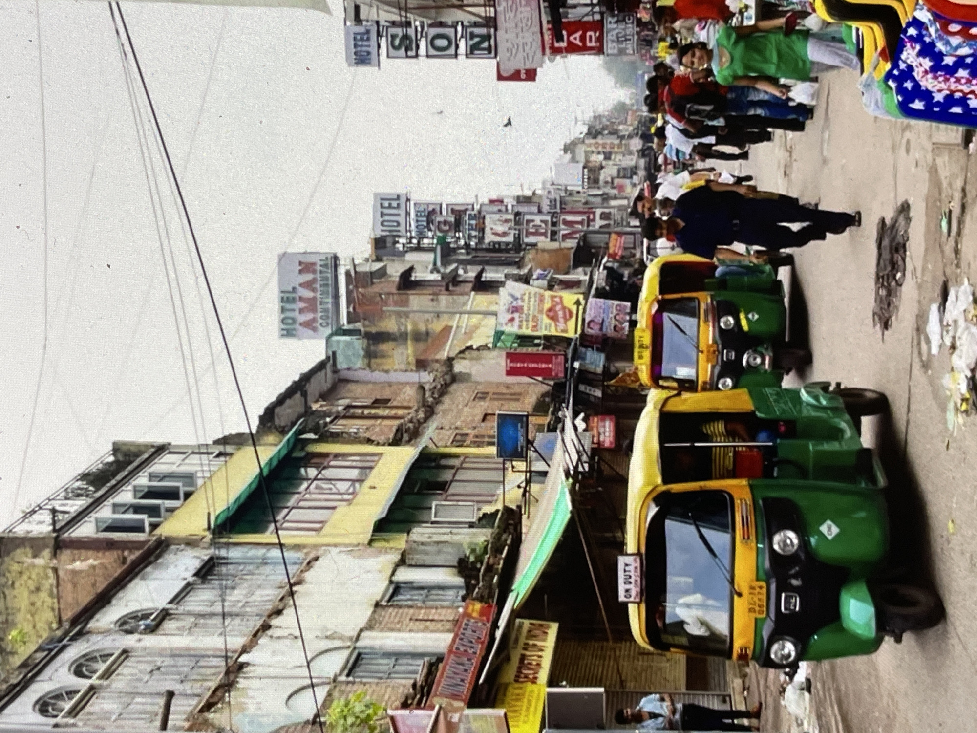 A street scene in what appears to be an Indian city. The image is dominated by buildings on either side of a busy street, filled with people and vehicles. The architecture suggests an older, possibly crowded urban environment, with multiple stories, balconies, and signs in a non-English script.
In the foreground, a green and yellow auto-rickshaw, a common mode of transport in South Asia, is prominently displayed. It has "ON DUTY" signage on its roof. Another similar auto-rickshaw is visible further down the street.
People are walking on the street, some appearing to be shoppers or pedestrians. One woman in a bright green top is walking towards the camera, carrying a white bag. A man in a dark blue shirt and pants is walking away from the camera.
Several signs for hotels are visible, including "HOTEL AMAN CONTINENTAL" and "HOTEL SON". Other signs, likely for businesses, are also present. The street is somewhat littered, with debris visible on the ground. Power lines and cables crisscross the sky. The overall impression is one of a bustling, active street in a densely populated area.