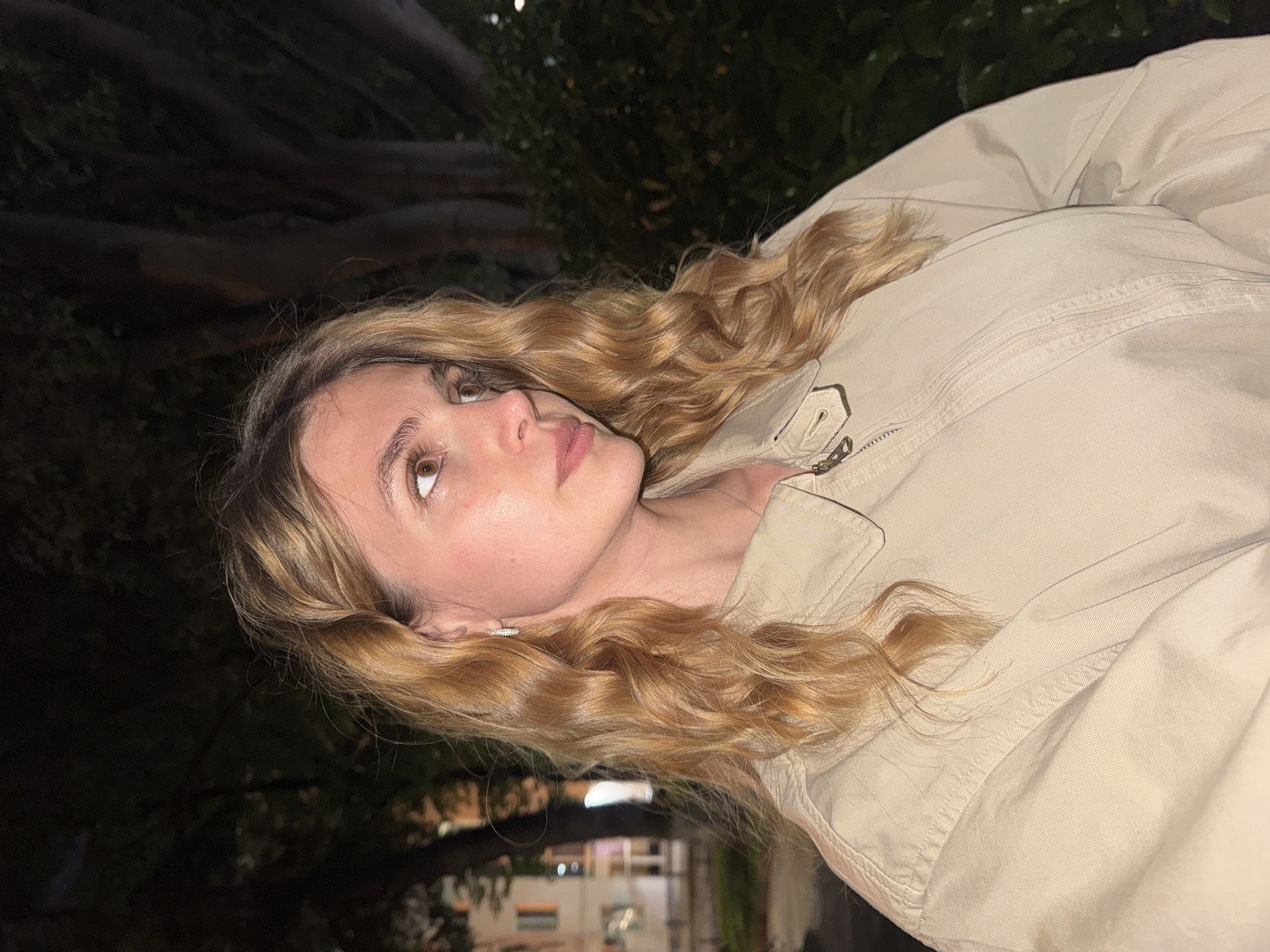A young woman with light brown hair is pictured wearing a beige jacket. She is outdoors at night, likely in Tbilisi, Georgia, based on the architectural style of buildings visible in the blurred background. The background shows foliage and dimly lit buildings. The focus is on the woman, who is looking upwards.