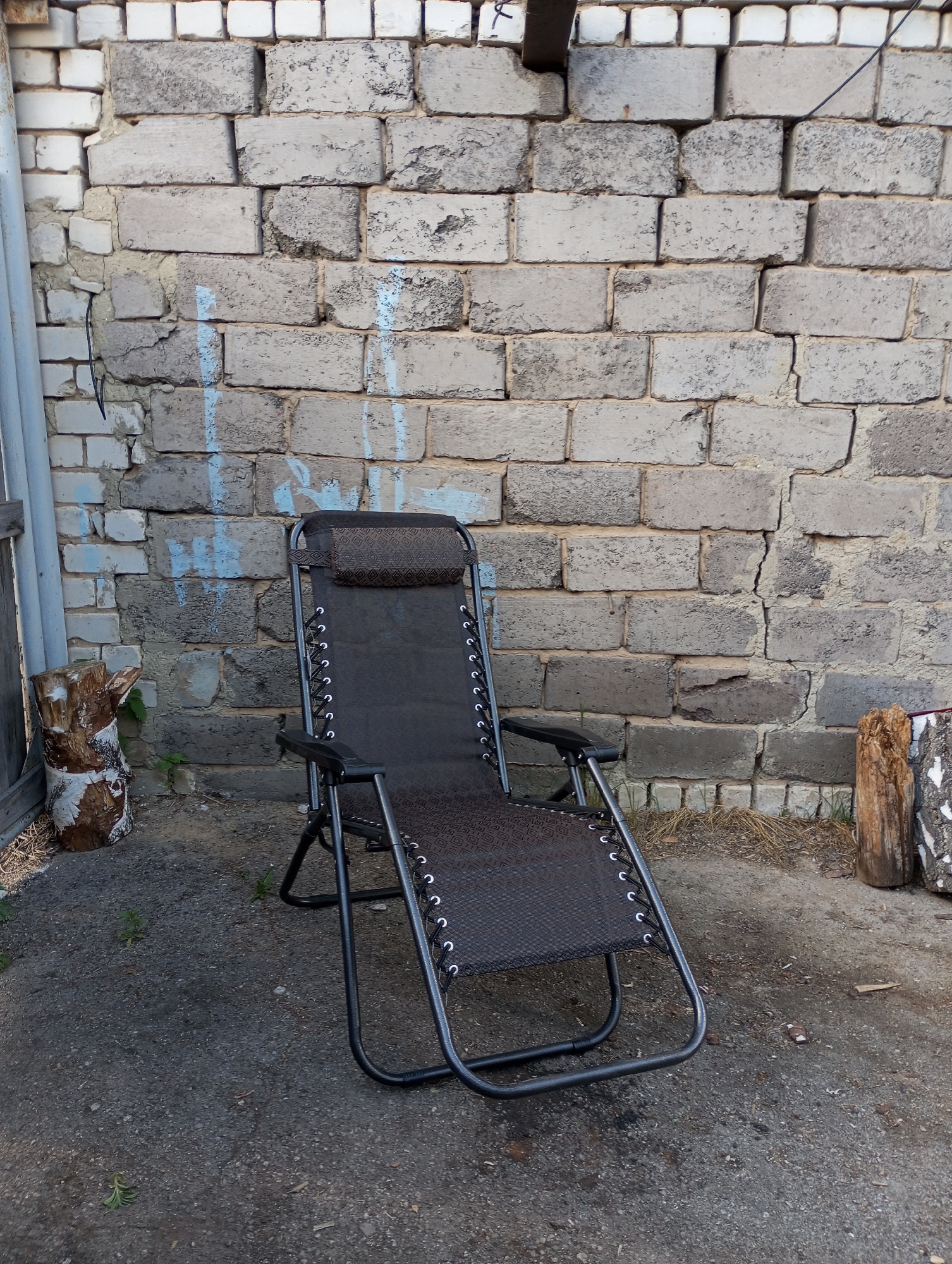 A dark-colored reclining chair stands on a paved surface in Кодинка, Russia. The chair is positioned in front of a light grey brick wall with some minor blue graffiti markings. To the left and right of the chair, at the base of the wall, are sections of cut wood. A partial white frame is visible on the left side of the image.