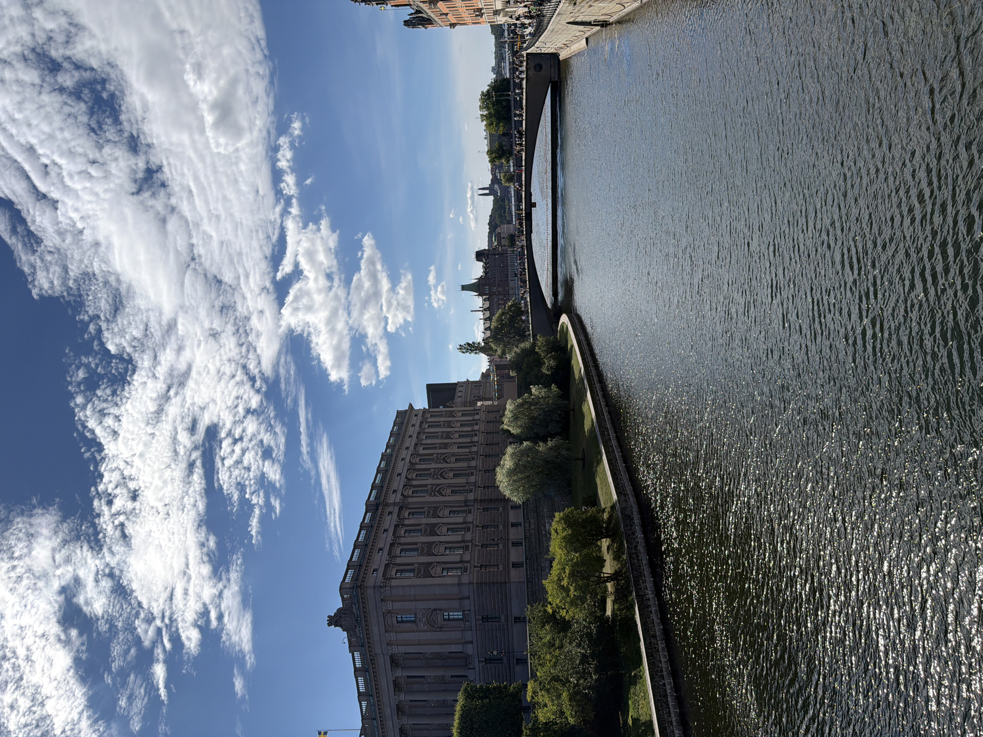 This is a photograph of the Riksdag building in Stockholm, Sweden, taken from a bridge over the Riksgatan waterway. The Riksdag building, also known as the Parliament House, is a large, neoclassical building with a prominent facade and a Swedish flag flying from its roof. The waterway is calm with a slight ripple, reflecting the bright sunlight. On the left bank of the waterway, there is a manicured lawn with several trees. On the right bank, there is a stone embankment and a multi-story building with an orange facade. A bridge spans the waterway in the middle ground, with numerous people walking across it. In the background, the Stockholm skyline is visible, with various buildings and structures under a blue sky with scattered white clouds. The overall scene depicts a sunny day in a European city, with a focus on the architectural landmarks and the waterway.